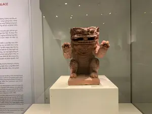 Bối cảnh là một không gian trưng bày trong bảo tàng hoặc một cuộc triển lãm. Ở trung tâm là một hiện vật khảo cổ được đặt trên bệ trắng và bảo vệ trong tủ kính. Hiện vật là một bức tượng linh vật bằng đất nung, có thể là con Nghê hoặc sư tử, trong tư thế ngồi với hai chân trước giơ lên và miệng mở. Bên trái hiện vật là một bảng thông tin lớn có chú thích bằng tiếng Việt và tiếng Anh. Nội dung văn bản đề cập đến lịch sử và kiến trúc của Hoàng thành Thăng Long. Một phần nội dung tiếng Việt có thể đọc được như sau: "...đứng, là khu vực đã tìm... cùng tường bao, cống... cung Thăng Long qua... ng về lịch sử phát triển... hộ phủ gắn với tên gọi... gia Đại Việt. Từ mùa Thu... (nghĩa là Rồng bay lên),... (1225-1400) và Hậu Lê... diễn ra tại đây vào năm... của các kiến trúc cung... tại đến ngày nay, nhưng đặc biệt là các loại ngói lợp... ơng, các loại ngói trang trí... à hình ảnh phản chiếu về... ược thiết kế rất công phu, quyền và thần quyền, thể... ây sẽ cho chúng ta những... a kiến trúc cung điện Việt... ắc thái độc đáo, riêng biệt". Ánh sáng từ đèn trên trần nhà phản chiếu lên mặt kính của tủ trưng bày.。（日本語訳）。背景は博物館または展覧会の展示スペースです。中央には、白い台座の上に置かれ、ガラスケースで保護された考古学的遺物があります。遺物はテラコッタ製の霊獣の像で、おそらくゲ（Nghê）または獅子であり、前足を上げて口を開けた座った姿勢をしています。遺物の左側には、ベトナム語と英語のキャプションが付いた大きな案内板があります。文書の内容は、タンロン皇城の歴史と建築に言及しています。ベトナム語の内容の一部は次のように読み取れます。「...立っており、発見された区域である...囲壁、排水溝と共に...タンロン宮殿を...通して...発展の歴史について...大越国の...という名に関連する官庁。秋から...（昇龍を意味する）、...（1225-1400）と後黎朝...が...年にここで開催された...宮殿建築の...今日まで存在するが、特に屋根瓦の種類...、装飾瓦の種類...についての反映されたイメージである...非常に精巧に設計され、権力と神権、体...これは我々に...ベトナムの宮殿建築の...独特で、固有の色彩を...与えるだろう」。天井の照明からの光が、展示ケースのガラス面に反射しています。