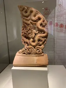 Đây là hình ảnh một hiện vật khảo cổ được trưng bày trong bảo tàng hoặc phòng triển lãm. Hiện vật chính là một tác phẩm điêu khắc bằng đất nung, có thể là một phần của đồ trang trí kiến trúc trên mái của một công trình cổ, chẳng hạn như Hoàng thành Thăng Long. Tác phẩm miêu tả một con rồng được chạm khắc vô cùng tinh xảo và sống động, với thân mình uốn lượn và các chi tiết phức tạp ở đầu và bờm. Hiện vật này bị vỡ một phần, được đặt trên một bệ đỡ màu trắng và bảo vệ trong một tủ kính. Phía sau tủ kính là một tấm phông nền có hình vẽ minh họa bằng đường nét màu đỏ, phác họa lại cấu trúc mái ngói và các linh vật trang trí khác, giúp người xem hình dung về vị trí ban đầu của hiện vật. Không có chữ viết nào trong ảnh.。（日本語訳）。これは、博物館または展示室に展示されている考古学的遺物の写真です。主な遺物はテラコッタ（素焼き）の彫刻作品で、タンロン皇城などの古代建築物の屋根の上にあった建築装飾の一部であった可能性があります。この作品は、非常に精巧かつ生き生きと彫刻された龍を描写しており、その体はうねり、頭とたてがみには複雑なディテールが施されています。この遺物は一部が破損しており、白い台座の上に置かれ、ガラスケースの中で保護されています。ガラスケースの後ろには、赤い線画のイラストが描かれた背景パネルがあり、瓦屋根の構造や他の装飾的な霊獣をスケッチして、鑑賞者が遺物の元の位置を想像するのに役立っています。写真には文字はありません。