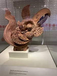 Bên trong một bảo tàng hoặc phòng trưng bày, một hiện vật khảo cổ được đặt trên bệ đỡ màu trắng và trong tủ kính. Đây là một tượng đầu chim phượng bằng đất nung, được chạm khắc tinh xảo với nhiều hoa văn phức tạp trên mào, mắt và cổ. Phía trước hiện vật có một bảng thông tin ghi rõ bằng tiếng Việt và tiếng Anh. Nội dung tiếng Việt là: "Tượng đầu chim phượng trang trí mái kiến trúc. Đất nung, thời Lý, thế kỷ 11-12". Phía sau tủ kính là một bức tường hoặc rèm có hoa văn trang trí.。（日本語訳）。博物館または展示室の内部、考古学的遺物が白い台座の上に置かれ、ガラスケースの中に収められています。これはテラコッタ製の鳳凰の頭部の像で、とさか、目、首に多くの複雑な文様が精巧に彫られています。遺物の前方には、ベトナム語と英語で明記された案内板があります。ベトナム語の内容は、「建築物の屋根を装飾する鳳凰の頭部の像。テラコッタ、李朝時代、11～12世紀」です。ガラスケースの背後には、装飾文様のある壁またはカーテンがあります。