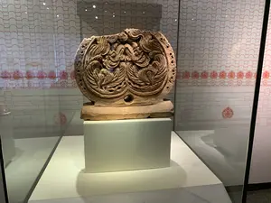 Bức ảnh chụp một hiện vật khảo cổ được trưng bày trong tủ kính tại một bảo tàng hoặc phòng triển lãm. Hiện vật chính là một tác phẩm điêu khắc bằng đất nung, có thể là một chi tiết trang trí kiến trúc, được đặt trên một bệ đỡ màu trắng. Tác phẩm điêu khắc mô tả một cặp phượng hoàng đối xứng nhau, được chạm khắc vô cùng tinh xảo với các đường nét uốn lượn phức tạp. Hiện vật bị vỡ ở phần trên và có một lỗ tròn ở phần đế. Phía sau tủ kính là một tấm phông nền có hoa văn trang trí màu đỏ trên nền trắng, mô phỏng hình ảnh ngói lợp và các họa tiết cổ. Đây là một hoạt động trưng bày hiện vật lịch sử, có thể liên quan đến khu di tích Hoàng thành Thăng Long.。（日本語訳）。この写真は、博物館または展示室のガラスケースの中に展示されている考古学的遺物を撮影したものです。主な遺物はテラコッタ製の彫刻作品で、建築装飾の一部であった可能性があり、白い台座の上に置かれています。この彫刻作品は、互いに対称な一対の鳳凰を描写しており、複雑で曲線的な線で非常に精巧に彫られています。遺物は上部が破損しており、基部には丸い穴があります。ガラスケースの後ろには、白地に赤の装飾文様が施された背景パネルがあり、屋根瓦や古代の文様を模しています。これは歴史的遺物の展示活動であり、タンロン皇城遺跡地区に関連している可能性があります。