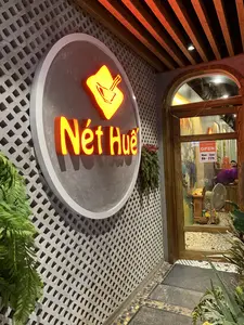 Hình ảnh chụp lối vào của một nhà hàng có tên 'Nét Huế'. Nổi bật là biển hiệu tròn lớn gắn trên tường, có chữ 'Nét Huế' và logo hình bát đũa được chiếu sáng bằng đèn neon màu cam. Bức tường bên cạnh được làm bằng gạch thông gió có hoa văn mắt cáo. Cửa ra vào làm bằng gỗ và kính, trên cửa có dán một biển báo nhỏ màu cam ghi 'Nét Huế', 'OPEN', và giờ hoạt động 'Mon - Sun 8h - 22h' (Thứ Hai - Chủ Nhật, 8 giờ sáng - 10 giờ tối). Nhìn qua cửa kính vào bên trong, có thể thấy một phần không gian nhà hàng với một người đang ngồi, một chiếc quạt điện, và một bàn thờ nhỏ. Lối đi và khu vực xung quanh được trang trí bằng cây xanh.。（日本語訳）。「Nét Huế」という名前のレストランの入り口を撮影した写真。目立つのは壁に取り付けられた大きな円形の看板で、「Nét Huế」の文字と、お椀と箸の形をしたロゴがオレンジ色のネオンで照らされている。隣の壁は、網目模様の透かしブロックでできている。出入り口のドアは木とガラスでできており、ドアには「Nét Huế」、「OPEN」、そして営業時間「月曜～日曜 午前8時～午後10時」と書かれたオレンジ色の小さな表示が貼られている。通路と周辺は緑の植物で飾られている。