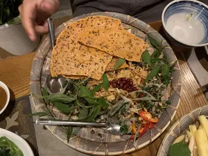 Hình ảnh chụp cận cảnh một món ăn Việt Nam được bày trên một chiếc mẹt tre. Món ăn chính có vẻ là hến xúc bánh tráng, một loại gỏi trộn. Các thành phần có thể nhận thấy bao gồm hến, rau răm, hành phi, đậu phộng rang, hành tây, và ớt tươi cắt lát. Phía trên món gỏi là những miếng bánh tráng nướng mè đen giòn. Có hai chiếc thìa inox được đặt trên mẹt để ăn. Xung quanh là các vật dụng khác trên bàn ăn như một bát nước chấm, một bát canh nhỏ, và có thể thấy một phần cánh tay của một người đang dùng bữa. Bối cảnh là một bữa ăn, có thể tại một nhà hàng hoặc tại nhà.。（日本語訳）。ベトナム料理一品を竹製の盆に盛り付けたクローズアップ写真です。メインの料理は、和え物の一種である「ヘン・スック・バインチャン」のようです。見て取れる具材には、シジミ、ベトナムコリアンダー、フライドオニオン、ローストピーナッツ、玉ねぎ、そしてスライスされた生の唐辛子が含まれています。和え物の上には、カリカリの黒ゴマ入り焼きライスペーパーが乗っています。盆の上には、食べるためのステンレス製のスプーンが2本置かれています。周りには、つけダレの椀、小さなスープの椀といった食卓の上の他の物があり、食事をしている人の腕の一部が見えます。背景は食事の場面で、レストランか自宅での食事だと思われます。