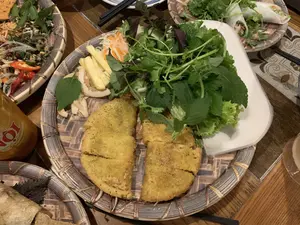 Trên nhãn chai bia có chữ "NỘI", là một phần của logo thương hiệu Bia Hà Nội. Đây là hình ảnh một bữa ăn với các món ăn Việt Nam được bày trên bàn gỗ. Món ăn nổi bật nhất là một đĩa bánh xèo được đặt trong mẹt tre. Bánh xèo màu vàng, giòn, được cắt thành nhiều miếng. Đi kèm với bánh xèo là một đĩa rau sống rất phong phú gồm xà lách, tía tô, rau mùi và các loại rau thơm khác, cùng với dứa thái lát và đồ chua (cà rốt, củ cải thái sợi). Trong khung hình còn có các món ăn khác như gỏi cuốn và một đĩa nộm ở phía sau. Bối cảnh cho thấy một bữa ăn tại nhà hàng hoặc một buổi sum họp gia đình.。（日本語訳）。ビールの瓶のラベルには、ハノイビールのブランドロゴの一部である「NỘI」という文字がある。これは、木製のテーブルに並べられたベトナム料理の食事の画像である。最も目立つ料理は、竹製の盆に置かれた一皿のバインセオである。バインセオは黄色でパリパリしており、いくつかのピースにカットされている。バインセオに添えられているのは、レタス、シソ、コリアンダー、その他の香草を含む非常に豊富な生野菜の皿と、スライスしたパイナップル、そして漬物（千切りにした人参、大根）である。フレームの中には、生春巻きや後ろにある一皿の和え物のような他の料理もある。背景は、レストランでの食事か、家族の集まりであることを示している。