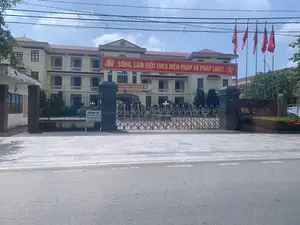 Bức ảnh chụp mặt tiền của một tòa nhà hành chính công, là trụ sở của một cơ quan chính quyền cấp huyện. Tòa nhà có kiến trúc cổ điển, sơn màu vàng, mái ngói đỏ, và có Quốc huy Việt Nam ở mặt tiền. Phía trước tòa nhà có treo một băng rôn lớn màu đỏ với khẩu hiệu 'SỐNG, LÀM VIỆC THEO HIẾN PHÁP VÀ PHÁP LUẬT。'. Một băng rôn khác phía trên cổng chính có dòng chữ '...XÃ HỘI CHỦ NGHĨA VIỆT NAM MUÔN NĂM。'. Bên phải là một hàng cột cờ treo nhiều lá cờ đỏ sao vàng của Việt Nam. Lối vào chính được đóng bằng một cổng xếp bằng kim loại. Gần cổng có một biển báo màu trắng ghi rõ quy định: 'XUỐNG XE, XUẤT TRÌNH GIẤY TỜ. KHÔNG MUA, BÁN HÀNG TRONG CƠ QUAN.'. Một biển hiệu lớn màu nâu ở cổng ghi 'TRỤ SỞ HĐND - UBND HUYỆN...' và 'ĐỊA CHỈ: KHU...'. Khung cảnh chung là một cảnh tĩnh, trang trọng tại một cơ quan nhà nước, không có hoạt động của người dân được ghi lại.。（日本語訳）。公共行政庁舎の正面を撮影した写真で、郡レベルの政府機関の本部である。建物は古典的な建築様式で、黄色に塗られ、赤い瓦屋根で、正面にはベトナムの国章がある。建物の前には「憲法と法律に従って生き、働く。正門の上にある別の横断幕には「...ベトナム社会主義共和国万歳。」という文字がある。右側には、ベトナムの金星紅旗が多数掲げられた旗竿が並んでいる。正面玄関は金属製の伸縮門で閉められている。門の近くには、「降車し、身分証明書を提示すること。庁舎内での物品の売買は禁止。」という規則を明記した白い看板がある。門にある大きな茶色の看板には、「...郡人民評議会・人民委員会本部」および「住所：...区」と書かれている。全体的な光景は、国家機関における静かで荘厳な場面であり、市民の活動は記録されていない。