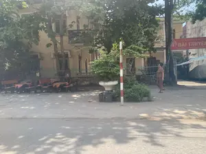 Ghi nhận chữ viết trên biển hiệu màu đỏ: 'ỦY BAN NHÂN DÂN HUYỆN... ĐÀI TRUYỀN THANH. ĐỊA CHỈ: KHU 3 - THỊ TRẤN VĨNH LỘC...'. Một phần chữ bị che khuất.。Bối cảnh là một con đường có vỉa hè rợp bóng cây tại một thị trấn. Một người phụ nữ lớn tuổi trong trang phục mặc nhà đang đi bộ trên vỉa hè. Phía sau là một tòa nhà hai tầng cũ, có thể là một cơ quan công quyền dựa trên biển hiệu. Dọc vỉa hè có nhiều băng ghế dài và một vài bộ bàn ghế nhỏ, có thể là của một quán nước. Ngoài ra còn có một cột mốc sơn sọc đỏ trắng, một chậu cây cảnh lớn, và một chiếc xe hơi được che bạt ở phía xa.。（日本語訳）。赤い看板の文字を記録：「…県人民委員会…放送局。住所：ヴィンロック町第3区…」。文字の一部は隠れている。背景は、ある町の並木道の歩道である。普段着の高齢女性が歩道を歩いている。後ろには古い2階建ての建物があり、看板から察するに公共機関の可能性がある。歩道沿いには多くの長いベンチといくつかの小さなテーブルと椅子のセットがあり、おそらく喫茶店のものであろう。その他、赤と白の縞模様に塗られた標柱、大きな観葉植物の鉢、そして遠くにシートで覆われた車が一台ある。