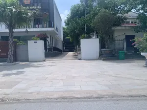 Bức ảnh chụp quang cảnh ban ngày tại một khu vực cơ quan hành chính. Có hai tòa nhà chính. Tòa nhà bên trái là một công trình hiện đại, trên đó có biển hiệu "VIỆN KIỂM SÁT NHÂN DÂN TỈNH THANH HÓA" và có gắn quốc huy Việt Nam. Tòa nhà bên phải, một phần bị cây cối che khuất, có biển hiệu "TÒA ÁN NHÂN DÂN HUYỆN VĨNH LỘC". Lối vào giữa hai tòa nhà là một con dốc dẫn vào bãi đậu xe bên trong, có thể thấy vài chiếc ô tô. Một cột cổng màu trắng có gắn biển số nhà "139". Phía trước là vỉa hè rộng được lát gạch đá. Xung quanh có nhiều cây xanh, bao gồm cây cọ và giàn hoa giấy. Một thùng rác màu xanh lá cây được đặt ở bên phải. Khung cảnh vắng lặng, không có hoạt động của con người.。（日本語訳）。ある行政機関エリアの日中の光景を撮影した写真です。主な建物が2つあります。左側の建物は近代的な建造物で、「VIỆN KIỂM SÁT NHÂN DÂN TỈNH THANH HÓA」（タインホア省人民検察院）という看板があり、ベトナムの国章が取り付けられています。右側の建物は、一部が木々で隠れていますが、「TÒA ÁN NHÂN DÂN HUYỆN VĨNH LỘC」（ヴィンロック県人民裁判所）という看板があります。2つの建物の間の入口は、中の駐車場へと続く坂道になっており、数台の車が見えます。白い門柱には「139」という番地プレートが取り付けられています。手前には石のタイルで舗装された広い歩道があります。周囲にはヤシの木やブーゲンビリアの棚など、多くの緑の木々があります。右側には緑色のゴミ箱が置かれています。辺りは静まり返っており、人の活動は見られません。