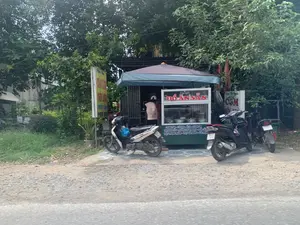 Đây là một quán ăn nhỏ ven đường, có một quầy hàng di động bán đồ ăn sẵn. Trên quầy hàng có biển hiệu lớn ghi 'BÁN ĐỒ ĂN SẴN' và liệt kê thực đơn gồm nhiều món ăn Việt Nam như: 'CANH CUA - CÁ KHO - ẾCH - LƯƠN OM', 'THỊT KHO TÀU - SƯỜN CHUA NGỌT - GIẢ CẦY', 'THỊT TRỨNG MẮM - THỊT GÀ RANG - GÀ TRỘN'. Bên cạnh đó còn có các biển hiệu khác quảng cáo 'CƠM', 'BÚN', và một biển hiệu màu vàng ghi 'CHÁO LƯƠN', 'MIẾN LƯƠN', 'PHỞ BÒ'. Một người phụ nữ đang đứng mua hàng tại quầy. Phía trước quán có một vài chiếc xe máy đang đậu. Khung cảnh xung quanh có nhiều cây xanh.。（日本語訳）。これは道端の小さな食堂で、惣菜を売る移動式の屋台があります。屋台には「BÁN ĐỒ ĂN SẴN」（惣菜販売）と書かれた大きな看板があり、「CANH CUA（カニのスープ） - CÁ KHO（魚の煮付け） - ẾCH（カエル） - LƯƠN OM（ウナギの煮込み）」、「THỊT KHO TÀU（豚の角煮） - SƯỜN CHUA NGỌT（甘酢スペアリブ） - GIẢ CẦY（犬肉風煮込み）」、「THỊT TRỨNG MẮM（豚肉と卵の魚醤煮） - THỊT GÀ RANG（鶏肉の炒め物） - GÀ TRỘN（鶏肉の和え物）」といった多くのベトナム料理のメニューが記載されています。その隣には「CƠM」（ご飯）、「BÚN」（ブン）を宣伝する他の看板もあり、黄色い看板には「CHÁO LƯƠN」（ウナギのお粥）、「MIẾN LƯƠN」（ウナギの春雨）、「PHỞ BÒ」（牛肉のフォー）と書かれています。一人の女性が屋台で買い物をしています。店の前には数台のバイクが停まっています。周囲の景色には緑の木々が多く見られます。