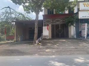 Bối cảnh là một con đường tại một thị trấn ở Việt Nam, có nhà dân và cửa hàng kinh doanh. Các văn bản có thể nhận dạng được bao gồm:。- Trên các biển hiệu của một cửa hàng bên phải: "TÓC", số điện thoại "088 512 1.9", "GỘI ĐẦU - NAIL", và địa chỉ "8 NGÃ TƯ HÀ LƯƠNG - THỊ TRẤN...". Một biển hiệu đứng khác quảng cáo "TÓC NAM ĐẸP", "CẮT TÓC NAM", phục vụ "24/24h" và các dịch vụ khác.。- Số nhà "171" được gắn trên tường một ngôi nhà.。- Bên trái có một tấm biển màu đỏ đã phai màu, chữ viết không thể đọc rõ.。Về quang cảnh, có một ngôi nhà với cổng sắt lớn màu xám, phía trước treo một lá cờ đỏ. Bên cạnh là một tiệm làm tóc, phía trước có một chiếc xe máy đang đậu. Ven đường có cây xanh lớn. Con đường được trải nhựa. Không có hoạt động rõ ràng của con người, nhưng khung cảnh cho thấy đây là một khu vực dân cư và có hoạt động thương mại.。（日本語訳）。背景はベトナムのある町の通りで、民家や商店があります。識別可能なテキストには以下が含まれます：。- 右側の店の看板には、「TÓC」、電話番号「088 512 1.9」、「GỘI ĐẦU - NAIL」、そして住所「8 NGÃ TƯ HÀ LƯƠNG - THỊ TRẤN...」。別の立て看板は「TÓC NAM ĐẸP」、「CẮT TÓC NAM」を広告し、「24/24h」対応やその他のサービスを宣伝しています。- 家屋番号「171」が家の壁に取り付けられています。- 左側には色褪せた赤い看板があり、文字ははっきりと読み取れません。風景については、大きな灰色の鉄の門がある家があり、その前には赤い旗が掲げられています。隣は美容院で、その前にはバイクが一台停まっています。道端には大きな緑の木があります。道路は舗装されています。はっきりとした人の活動は見られませんが、この風景はここが住宅地であり、商業活動があることを示しています。