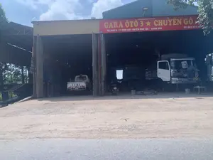 Hình ảnh cho thấy mặt tiền của một gara sửa chữa ô tô. Phía trên cửa ra vào có một tấm biển hiệu lớn màu đỏ, chữ vàng ghi 'GARA ÔTÔ 3 ★ CHUYÊN GÒ'. Dưới đó là địa chỉ 'ĐC: KHU 5 - TT VĨNH LỘC - HUYỆN VĨNH LỘC - THANH HÓA' và số điện thoại 'ĐT: 0915 872 569'. Có một biển số nhà nhỏ màu xanh ghi số '293'.。Đây là một xưởng sửa chữa xe cộ. Bên trong gara có không gian rộng, có nhiều xe tải đang trong quá trình sửa chữa. Một người thợ đang đứng sửa chữa phần đầu của một chiếc xe tải màu trắng ở bên phải. Một chiếc xe tải nhỏ màu trắng khác, có chữ 'TTDT' ở phía sau, đang đỗ ở bên trái. Ngoài ra còn có một chiếc xe tải màu sẫm và một chiếc xe máy ở bên trong. Sân trước gara là nền đất.。（日本語訳）。写真は自動車修理工場の正面を写しています。入り口の上には大きな赤い看板があり、黄色の文字で「GARA ÔTÔ 3 ★ CHUYÊN GÒ」と書かれています。その下には住所「ĐC: KHU 5 - TT VĨNH LỘC - HUYỆN VĨNH LỘC - THANH HÓA」と電話番号「ĐT: 0915 872 569」が記載されています。「293」と書かれた小さな青い番地プレートがあります。ここは車両の修理工場です。ガレージの中は広い空間で、多くのトラックが修理中です。右側では、一人の整備士が白いトラックのフロント部分を立って修理しています。後部に「TTDT」という文字がある別の白い小型トラックが、左側に駐車しています。その他にも、中には濃い色のトラック1台とバイク1台があります。ガレージの前の庭は土の地面です。