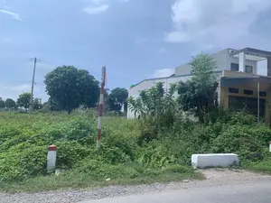 Bức ảnh chụp một khung cảnh ven đường ở khu vực nông thôn hoặc ngoại ô. Bên phải là một ngôi nhà hiện đại, nhiều tầng, sơn màu trắng và be, có ban công. Bên cạnh ngôi nhà là một khu đất trống mọc um tùm cây cỏ dại. Dọc theo lề đường có các cọc tiêu giao thông sơn sọc đỏ trắng và một khối bê tông màu trắng. Ở phía sau, có thể thấy nhiều cây xanh lớn và một cột điện với đường dây điện. Bầu trời trong xanh với vài gợn mây trắng. Không có người hay hoạt động nào đang diễn ra. Có một biển báo gắn trên cọc tiêu nhưng không thể đọc được nội dung do góc chụp.。（日本語訳）。写真は、田舎または郊外の道端の風景を写しています。右側には、白とベージュ色に塗られた、バルコニー付きの近代的な多層階の家があります。家の隣には、雑草が生い茂る空き地があります。道端に沿って、赤と白の縞模様に塗られた交通用の杭と、白いコンクリートブロックがあります。後方には、多くの大きな緑の木々と、電線のある電柱が見えます。空は青く澄んでおり、白い雲がいくつか浮かんでいます。人や活動は見られません。杭には標識が取り付けられていますが、撮影角度のため内容は読み取れません。