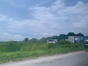 Bức ảnh chụp một khung cảnh ven đường ở vùng nông thôn. Bên trái là một cánh đồng lúa xanh tươi, phía xa là những dãy núi đá. Ở giữa bức ảnh là một tòa nhà lớn có mái tôn màu xanh, có thể là một nhà kho hoặc cửa hàng vật liệu xây dựng. Một chiếc xe tải nhỏ màu trắng đang đỗ gần tòa nhà. Có một tấm biển quảng cáo lớn màu xanh dương, trên đó có các dòng chữ: "NHÀ PHÂN PHỐI LONG HƯƠNG", logo "JYMEC", "SƠN & CHỐNG THẤM", "ĐỒ ĐIỆN", "THIẾT BỊ VỆ SINH - M...". Bên phải, một người đang lái xe máy trên con đường nhựa. Bầu trời có nhiều mây.。（日本語訳）。この写真は、田舎の道端の風景を写したものです。左側には青々とした水田が広がり、遠くには岩山が連なっています。写真の中央には、青いトタン屋根の大きな建物があります。倉庫か建材店のようです。建物の近くに白い小型トラックが1台停まっています。大きな青い看板があり、そこには「NHÀ PHÂN PHỐI LONG HƯƠNG」、「JYMEC」のロゴ、「SƠN & CHỐNG THẤM」、「ĐỒ ĐIỆN」、「THIẾT BỊ VỆ SINH - M...」という文字が書かれています。右側では、一人の人がアスファルトの道路をバイクで走っています。空には雲が多く浮かんでいます。