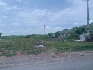 Bức ảnh chụp một khung cảnh vùng nông thôn. Ở trung tâm bức ảnh là một con trâu đang đứng trên bãi cỏ. Phía sau con trâu là một cánh đồng rộng lớn, xa hơn là những ngọn đồi núi trập trùng. Bầu trời trong xanh và có nhiều mây trắng. Có các cột điện và đường dây điện chạy ngang qua khung cảnh. Ở bên phải, gần đường, có một số khối bê tông, tấm lợp fibro xi măng xếp chồng lên nhau và một vài cây chuối. Mặt đất ở tiền cảnh là một bãi đất trống có cỏ mọc lưa thưa, sỏi đá và một vũng nước nhỏ. Không có văn bản hay chữ viết nào trong ảnh.。（日本語訳）。写真は田舎の風景を写しています。写真の中央には、草地に立っている一頭の水牛がいます。水牛の後ろには広大な野原が広がり、さらにその奥には起伏のある丘や山々があります。空は青く澄んでおり、白い雲がたくさん浮かんでいます。電柱と電線が風景を横切っています。右側、道路の近くには、いくつかのコンクリートブロック、積み重ねられた繊維セメントの屋根板、そして数本のバナナの木があります。前景の地面は、草がまばらに生え、砂利や石、そして小さな水たまりがある空き地です。写真にはテキストや文字はありません。