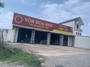 Hình ảnh cho thấy mặt tiền của một cửa hàng vật liệu xây dựng nằm ven đường. Cửa hàng có một tấm biển hiệu lớn màu đỏ với chữ vàng ghi 'TÔN HOA SEN' và khẩu hiệu 'Mái ấm gia đình Việt'. Một tấm biển nhỏ hơn ghi tên cửa hàng là 'CỬA HÀNG VĨNH LỘC', số điện thoại 'ĐT: 0237 352 0088' và địa chỉ 'Quốc Lộ 45, Thôn Thọ Vực 2, X. Ninh Khang, H. Vĩnh Lộc, T. Thanh Hóa'. Tòa nhà là một nhà kho lớn với nhiều cửa cuốn đang mở rộng. Bên trong các gian hàng, có nhiều vật liệu xây dựng được xếp chồng lên nhau, bao gồm các cuộn tôn hoặc lưới thép và các loại ống. Có một người đang làm việc ở phía trong một gian hàng. Bên cạnh nhà kho là một ngôi nhà dân cao tầng. Bầu trời có mây.。（日本語訳）。写真は、道端にある建材店の正面を写している。店には、「TÔN HOA SEN」と「Mái ấm gia đình Việt（ベトナムの家庭の暖かい家）」というスローガンが黄色い文字で書かれた大きな赤い看板がある。より小さな看板には、店名「CỬA HÀNG VĨNH LỘC（ヴィンロック店）」、電話番号「ĐT: 0237 352 0088」、住所「Quốc Lộ 45, Thôn Thọ Vực 2, X. Ninh Khang, H. Vĩnh Lộc, T. Thanh Hóa（タインホア省ヴィンロック県ニンカン社トーヴック2村、国道45号線）」が記載されている。建物は大きな倉庫で、多くのシャッタードアが大きく開いている。各区画の内側には、トタンや金網のロール、各種パイプなど、多くの建材が積み重ねられている。ある区画の内側で一人の人が作業している。倉庫の隣には、高層の民家がある。空は曇っている。