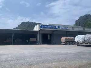 Hình ảnh ghi lại quang cảnh tại một nhà kho hoặc khu vực phân phối vật liệu xây dựng. Trên tòa nhà có hai biển hiệu. Biển hiệu màu xanh có logo 'KYOEI', dòng chữ 'THÉP KYOEI JAPAN', 'Công ty Nhật - Chất lượng Nhật' và hình lá cờ Nhật Bản. Biển hiệu màu trắng bên cạnh ghi 'CÔNG TY TNHH XIIK QUYẾT CƯỜNG'. Bối cảnh phía sau là những dãy núi đá vôi. Trong sân rộng rải sỏi, có nhiều xe tải hạng nặng đang đỗ. Một chiếc xe tải được chất đầy các bao tải, trong khi một chiếc khác được phủ bạt. Dưới một mái che, có thêm hai thùng xe tải cũng đang chất hàng. Hoạt động chính dường như là việc tập kết và vận chuyển hàng hóa. Có một vài chiếc xe máy đang đỗ gần lối vào nhà kho.。（日本語訳）。画像は、ある倉庫または建設資材の配送エリアの光景を記録したものです。建物には2つの看板があります。青い看板には、「KYOEI」のロゴ、「THÉP KYOEI JAPAN」、「Công ty Nhật - Chất lượng Nhật（日本の会社 - 日本の品質）」という文字、そして日本の国旗の絵があります。隣の白い看板には「CÔNG TY TNHH XIIK QUYẾT CƯỜNG」と書かれています。背景には石灰岩の山々が連なっています。砂利が敷かれた広い庭には、多くの大型トラックが駐車しています。1台のトラックには袋が満載されており、もう1台は防水シートで覆われています。屋根の下では、さらに2台のトラックの荷台にも荷物が積まれています。主な活動は商品の集荷と輸送のようです。倉庫の入り口近くに数台のバイクが停まっています。