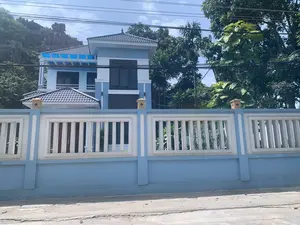 Bức ảnh chụp một ngôi nhà hiện đại, có thể là một biệt thự, nằm bên cạnh một con đường. Ngôi nhà được sơn màu xanh da trời và trắng, có kiến trúc nhiều tầng và mái lợp ngói màu sẫm. Phía trước là một hàng rào bê tông kiên cố, được sơn đồng màu với ngôi nhà. Ở hậu cảnh, có một ngọn núi đá vôi với cây cối bao phủ. Nhiều loại cây xanh tươi tốt mọc xung quanh khu vực nhà. Bầu trời trong xanh và có nhiều đường dây điện, dây cáp vắt ngang qua phía trên. Không có người hay hoạt động nào được ghi lại trong ảnh.。（日本語訳）。その家は水色と白で塗装されており、多層構造で、屋根は濃い色の瓦で葺かれています。正面には頑丈なコンクリートの塀があり、家と同じ色で塗装されています。背景には、木々に覆われた石灰岩の山があります。家の周りには、青々とした多くの種類の植物が生い茂っています。空は青く澄んでおり、上部には多くの電線やケーブルが横切っています。写真には人や活動は記録されていません。