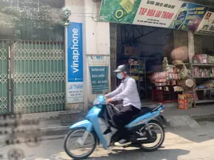 Bối cảnh là một con đường ở Việt Nam, phía trước một cửa hàng tạp hóa. Một người đàn ông đội mũ bảo hiểm và đeo khẩu trang đang điều khiển một chiếc xe máy màu xanh lam đang di chuyển trên đường. Bên cạnh là một cửa hàng tạp hóa có tên "TẠP HÓA LAI THANH", bên trong có các kệ trưng bày các mặt hàng như bánh kẹo, đồ ăn vặt. Phía trước cửa hàng có nhiều biển hiệu được ghi lại như sau:。1.  Biển hiệu chính của cửa hàng: "BIA RƯỢU - BÁNH KẸO - NƯỚC GIẢI KHÁT", "TẠP HÓA LAI THANH", "ĐT: 0981 083 586", "ĐC: QL45, NINH KHANG, VĨNH LỘC, THANH HÓA".。2.  Biển hiệu dọc màu xanh: "vinaphone", "VNPT".。3.  Biển hiệu nhỏ bên dưới biển Vinaphone: "LAI THANH", "ĐIỂM BÁN SIM - THẺ", "HÀNG TẠP HÓA".。4.  Băng rôn của công ty bảo hiểm VNI: "ĐIỂM BÁN BẢO HIỂM Ô TÔ - XE MÁY".。5.  Biển quảng cáo sản phẩm: "KHÔNG ĐỘ", "BÙ KHOÁNG", "TĂNG CƯỜNG SỨC MẠNH".。Bên cạnh cửa hàng tạp hóa là một ngôi nhà có cửa xếp bằng kim loại màu xanh lá cây.。（日本語訳）。背景はベトナムのある道路で、一軒の雑貨店の前です。ヘルメットをかぶりマスクを着けた男性が、青いバイクを運転して道路を走行しています。その隣には「TẠP HÓA LAI THANH」という名前の雑貨店があり、店内には菓子やスナック菓子などの商品が陳列された棚があります。店の前には多くの看板があり、以下のように記されています。1.  店のメイン看板：「ビール・酒 - 菓子 - 清涼飲料水」、「ライタイン雑貨店」、「電話：0981 083 586」、「住所：国道45号線、ニンカン社、ヴィンロック県、タインホア省」。2.  青い縦長の看板：「vinaphone」、「VNPT」。3.  Vinaphoneの看板の下にある小さな看板：「ライタイン」、「SIM・カード販売店」、「雑貨」。4.  VNI保険会社の横断幕：「自動車・バイク保険販売店」。5.  商品の広告看板：「ゼロ度」、「ミネラル補給」、「パワー増強」。雑貨店の隣には、緑色の金属製蛇腹式ドアのある家があります。