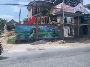 Bên cạnh một con đường nhựa là một công trình đang thi công cổng và tường rào của một ngôi nhà. Một người thợ xây đang làm việc trên giàn giáo làm bằng tre, phía trên có mái che tạm bằng bạt sọc. Dưới đất có các đống vật liệu xây dựng như cát và sỏi đá. Trên bức tường đang được xây dựng có vẽ hai bức tranh tường lớn mô tả phong cảnh thiên nhiên với núi non, sông nước, cây cối và một thác nước. Phía sau công trình là một ngôi nhà nhiều tầng có ban công, trên đó có treo một lá cờ đỏ. Ở phía bên phải của cổng đang xây, có một tấm biển màu xanh lá cây với chữ trắng ghi 'QUẦY THUỐC TÂN DƯỢC'.。（日本語訳）。アスファルトの道の隣に、ある家の門と塀の工事現場があります。一人の建設作業員が竹でできた足場の上で作業しており、その上には縞模様のシートでできた仮設の屋根があります。地面には砂や砂利などの建設資材の山があります。建設中の壁には、山、川、木々、そして滝のある自然の風景を描いた2つの大きな壁画が描かれています。工事現場の後ろにはバルコニーのある多層階の家があり、そこには赤い旗が掲げられています。建設中の門の右側には、「QUẦY THUỐC TÂN DƯỢC」と白文字で書かれた緑色の看板があります。