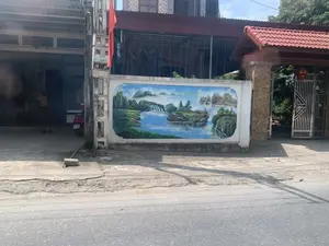 Hình ảnh chụp một con đường nhựa ở khu dân cư. Bên đường là một bức tường rào của một ngôi nhà, trên đó có vẽ một bức tranh phong cảnh sơn thủy hữu tình lớn với núi non, sông nước và thác nước. Cạnh đó là cổng nhà có cột trụ ốp đá, mái ngói đỏ và một chiếc đèn lồng đỏ treo ở cổng. Phía bên trái, có một cột điện bê tông, trên đó có treo một lá cờ đỏ và dán giấy quảng cáo với nội dung có thể đọc được là 'HÚT BỂ PHỐT' cùng các số điện thoại '0978 166...' và '0968 653.6...'. Bên trong hiên một ngôi nhà khác, có một chiếc xe máy màu đỏ đang đỗ.。（日本語訳）。住宅街のアスファルトの道を撮影した写真。道の脇にはある家の塀があり、その上には山、川、滝が描かれた大きな情趣あふれる山水風景画が描かれている。その隣には、石張りの柱、赤い瓦屋根、そして門に赤い提灯が吊るされた家の門がある。左側にはコンクリートの電柱があり、そこには赤い旗が掲げられ、「HÚT BỂ PHỐT」という内容と電話番号「0978 166...」および「0968 653.6...」が読み取れる広告紙が貼られている。別の家の軒の内側には、赤いバイクが1台停まっている。