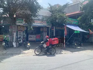 Bối cảnh là một con đường tại Việt Nam với nhiều cửa hàng, chủ yếu là các nhà thuốc, nằm san sát nhau bên vỉa hè. Ở trung tâm bức ảnh, một người giao hàng của Viettel Post trong bộ đồng phục màu đỏ đang ngồi trên xe máy. Phía sau xe có một thùng hàng lớn màu đỏ. Nhiều xe máy khác đang đậu trên vỉa hè. Cây xanh được trồng phía trước các cửa hàng.。Các thông tin từ các biển hiệu được ghi lại như sau:。1. Nhà thuốc ở trung tâm (Thành Dung Số 6):。- Biển hiệu chính: "NHÀ THUỐC PHẨM YÊN ĐỊNH", "THÀNH DUNG SỐ 6", "KINH DOANH DƯỢC PHẨM - THỰC PHẨM CHỨC NĂNG".。- Thông tin liên hệ: "GPĐK SỐ:", "ĐT: 0352 027 365".。- Quảng cáo: "Salonpas", "Hisamitsu", "HÀNG CHÍNH HÃNG CỦA HISAMITSU VIỆT NAM".。- Biển hiệu dọc: "QUẦY THUỐC ĐẠT", "BÁN VÀ TƯ VẤN HƯỚNG DẪN SỬ DỤNG THUỐC".。2. Nhà thuốc bên phải (Hằng Tỉnh):。- Biển hiệu chính: "CÔNG TY CỔ PHẦN DƯỢC - VTYT THANH HÓA", "NHÀ THUỐC LIÊN ANH DƯỢC PHẨM YÊN ĐỊNH", "QUẦY THUỐC HẰNG TỈNH".。- Thông tin chi tiết: "PHỤ TRÁCH CHUYÊN MÔN: DSĐH. DƯỢC SỸ PHẠM THỊ HẰNG", "Đ/C: SN 200 - QL.45 - Phố Kiều - Yên Trường - Yên Định - Thanh Hóa", "ĐT: 0904 606 244".。- Biển đèn LED: "HẰNG TỈNH".。3. Nhà thuốc bên trái:。- Biển hiệu: "QUẦY THUỐC HÀI", "BÁN LẺ THUỐC".。4. Thùng hàng của người giao hàng:。- Chữ trên thùng: "viettel post", "1900.8095".。（日本語訳）。背景はベトナムのある通りで、多くの店、主に薬局が歩道沿いに隣接して並んでいます。写真の中央には、赤い制服を着たViettel Postの配達員がバイクに座っています。バイクの後ろには、大きな赤い配達用の箱があります。他の多くのバイクが歩道に停まっています。店の前には緑の木々が植えられています。看板からの情報は以下の通りです：。1. 中央の薬局（Thành Dung Số 6）：。- メインの看板：「NHÀ THUỐC PHẨM YÊN ĐỊNH」（イエンディン薬局）、「THÀNH DUNG SỐ 6」（タインズン6号店）、「KINH DOANH DƯỢC PHẨM - THỰC PHẨM CHỨC NĂNG」（医薬品 - 機能性食品の販売）。- 連絡先情報：「GPĐK SỐ:」（登録許可番号：）、「ĐT: 0352 027 365」（電話：0352 027 365）。- 広告：「サロンパス」、「久光」、「HÀNG CHÍNH HÃNG CỦA HISAMITSU VIỆT NAM」（久光製薬ベトナムの正規品）。- 縦看板：「QUẦY THUỐC ĐẠT」（ダット薬局カウンター）、「BÁN VÀ TƯ VẤN HƯỚNG DẪN SỬ DỤNG THUỐC」（医薬品の販売および使用方法の相談・指導）。2. 右側の薬局（Hằng Tỉnh）：。- メインの看板：「CÔNG TY CỔ PHẦN DƯỢC - VTYT THANH HÓA」（タインホア製薬・医療機器株式会社）、「NHÀ THUỐC LIÊN ANH DƯỢC PHẨM YÊN ĐỊNH」（リエンアイン薬局 イエンディン医薬品）、「QUẦY THUỐC HẰNG TỈNH」（ハンティン薬局カウンター）。- 詳細情報：「PHỤ TRÁCH CHUYÊN MÔN: DSĐH. DƯỢC SỸ PHẠM THỊ HẰNG」（専門担当者：大学薬剤師 ファム・ティ・ハン）、「Đ/C: SN 200 - QL.45 - Phố Kiều - Yên Trường - Yên Định - Thanh Hóa」（住所：200番地 - 国道45号線 - キエウ通り - イエンチュオン - イエンディン - タインホア）、「ĐT: 0904 606 244」（電話：0904 606 244）。- LED看板：「HẰNG TỈNH」（ハンティン）。3. 左側の薬局：。- 看板：「QUẦY THUỐC HÀI」（ハイ薬局カウンター）、「BÁN LẺ THUỐC」（医薬品小売）。4. 配達員の配達用箱：。- 箱の文字：「viettel post」、「1900.8095」。