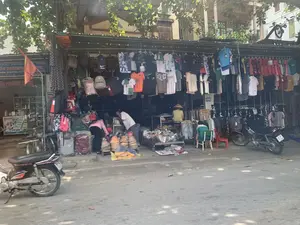 Bối cảnh là một cửa hàng ven đường ở Việt Nam, có thể là một khu chợ nhỏ. Dưới bóng cây xanh, một gian hàng được dựng lên trên vỉa hè, bày bán nhiều loại hàng hóa. Các mặt hàng chủ yếu là quần áo và ba lô, được treo san sát nhau trên các giá đỡ. Có rất nhiều áo phông, bao gồm cả các loại áo đấu thể thao, và các loại quần áo khác. Nhiều chiếc ba lô với màu sắc và kiểu dáng đa dạng cũng được treo bán. Dưới đất, có một chồng nón lá truyền thống của Việt Nam được bày bán. Một vài người đang ở gian hàng, có vẻ như đang lựa chọn và mua sắm. Một người phụ nữ mặc áo hồng đang cúi xuống xem hàng, một người đàn ông mặc áo trắng đang đứng xem, và một người khác đội nón lá đang ở sâu bên trong. Có hai chiếc xe máy đang đỗ gần đó. Bên trái gian hàng, có một cửa hàng khác với biển hiệu màu xanh ghi chữ "QUỐC PHƯƠNG THANH" và "DƯỢC PHẨM", cho thấy đó là một hiệu thuốc. Một lá cờ màu đỏ cũng được treo ở phía trước.。（日本語訳）。舞台はベトナムの道端にある店で、おそらく小さな市場でしょう。緑の木陰の下、歩道に露店が設けられ、様々な商品が並べられています。主な商品は衣類とバックパックで、ラックにぎっしりと掛けられています。スポーツユニフォームを含む多くのTシャツや、その他の衣類があります。様々な色やデザインの多くのバックパックも販売のために掛けられています。地面には、ベトナムの伝統的なノンラーが山積みになって売られています。数人が露店におり、商品を選んで買い物をしているようです。ピンクの服を着た女性がかがんで商品を見ており、白いシャツを着た男性が立って見ており、もう一人はノンラーをかぶって奥の方にいます。近くには2台のバイクが停まっています。露店の左側には、「QUỐC PHƯƠNG THANH」と「DƯỢC PHẨM」と書かれた青い看板の別の店があり、薬局であることがわかります。赤い旗も前面に掲げられています。