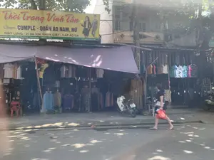 Bức ảnh chụp một cửa hàng quần áo ven đường có tên 'Thời Trang Vinh Tân'. Trên biển hiệu lớn màu vàng có ghi các thông tin: 'Chuyên COMPLE - QUẦN ÁO NAM, NỮ', 'DÂY LƯNG - VÍ DA XỊN. NHẬN GIẶT - HẤP ÁO DA', và địa chỉ 'PHỐ KIẾU - YÊN TRƯỜNG - YÊN ĐỊNH - THANH HÓA' cùng số điện thoại 'ĐT: 0934 643 829'.。Khung cảnh cho thấy một cửa hàng đang hoạt động kinh doanh. Rất nhiều quần áo được treo trên các giá đỡ ở phía trước, dưới một tấm bạt lớn màu tím được căng ra để che nắng. Bên trong cửa hàng, có một người phụ nữ đang ngồi trên chiếc ghế nhựa màu đỏ, và một chiếc xe máy đang đỗ. Trên con đường phía trước, một người phụ nữ khác đang đi bộ chân trần, tay xách một chiếc túi ni lông màu đỏ.。（日本語訳）。写真は「Thời Trang Vinh Tân」という名前の道端の衣料品店を写したものです。大きな黄色の看板には、「スーツ専門 - 紳士・婦人服」、「高級革ベルト - 革財布。革ジャンの洗濯・スチームクリーニング承ります」という情報と、「PHỐ KIẾU - YÊN TRƯỜNG - YÊN ĐỊNH - THANH HÓA」という住所、そして「電話：0934 643 829」という電話番号が書かれています。その光景は、店が営業中であることを示しています。たくさんの服が店の前のハンガーラックに掛けられており、日差しを遮るために張られた大きな紫色のタープの下にあります。店内には、赤いプラスチックの椅子に座っている女性が一人と、一台のバイクが停まっています。前の道では、別の女性が裸足で歩いており、手には赤いビニール袋を提げています。