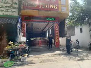 Bức ảnh chụp một con phố ở Việt Nam với các hoạt động thường ngày. Nổi bật là một tiệm vàng bạc lớn tên là "KIM TRUNG", có địa chỉ số 170. Biển hiệu chính ghi "TIỆM VÀNG BẠC KIM TRUNG" và một bảng điện tử LED đang chạy chữ "DOANH". Các biển hiệu dọc trên cột cũng ghi "VÀNG" và "KIM TRUNG". Bên cạnh là một cửa hàng có biển hiệu "Hồng", "XĂM VÀ ĐIỀU TRỊ DA", "đẹp của bạn" cùng thông tin liên hệ Facebook "Lê Hồng" và số điện thoại "0977 913 848". Trên vỉa hè, một người phụ nữ đội nón lá đang ngồi bán trái cây trên một chiếc xe đạp. Xe hàng của bà chất đầy dưa hấu và các loại quả khác, bên dưới có các sọt nhựa đựng thêm hoa quả và một rổ đậu bắp. Một người đàn ông mặc đồ đen đang đứng trên bậc thềm của tiệm vàng, trong khi một người đàn ông khác đang đứng cạnh chiếc xe đạp ở bên phải. Có một vài chiếc xe máy đang đỗ trên vỉa hè.。（日本語訳）。目立っているのは「KIM TRUNG」という名前の大きな金銀店で、住所は170番地です。メインの看板には「TIỆM VÀNG BẠC KIM TRUNG」と書かれており、LED電光掲示板には「DOANH」という文字が流れています。柱にある縦長の看板にも「VÀNG」と「KIM TRUNG」と書かれています。隣には、「Hồng」、「XĂM VÀ ĐIỀU TRỊ DA」、「đẹp của bạn」という看板を掲げた店があり、Facebookの連絡先「Lê Hồng」と電話番号「0977 913 848」が併記されています。歩道では、ノンラーをかぶった女性が自転車に座って果物を売っています。彼女の荷台にはスイカやその他の果物が満載で、その下にはさらに果物が入ったプラスチック製のカゴとオクラの入ったザルがあります。黒い服を着た男性が金店の階段に立っており、一方、別の男性が右側の自転車の隣に立っています。歩道には数台のバイクが停まっています。