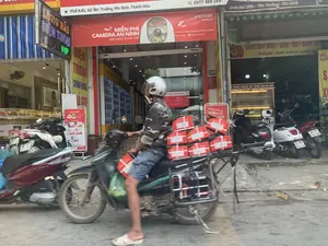 Bối cảnh là một con đường ở Phố Kiểu, Xã Yên Trường, Yên Định, Thanh Hóa. Ở trung tâm bức ảnh, một người đàn ông đang lái xe máy, đội mũ bảo hiểm, chở nhiều thùng hàng màu đỏ được buộc ở yên sau, có thể là một người giao hàng. Phía sau là mặt tiền của các cửa hàng. Có nhiều xe máy khác đang đậu trên vỉa hè. Các dòng chữ trên các biển hiệu có thể đọc được bao gồm: "Phố Kiểu, Xã Yên Trường, Yên Định, Thanh Hóa", "viettel telecom", "MIỄN PHÍ CAMERA AN NINH BẢO MẬT TUYỆT ĐỐI", "LONG HẢI ĐIỆN THOẠI SIM SỐ ĐẸP", "BÁNH SINH NHẬT - CƯỚI - HỎI", và một biển quảng cáo đứng của Viettel với nội dung "TẶNG MÁY 4G MIỄN PHÍ".。（日本語訳）。背景は、タインホア省イエンディン県イエンチュオン社キエウ通りのある道です。写真の中央では、一人の男性がヘルメットをかぶり、バイクを運転しており、後部座席に縛り付けられた多くの赤い箱を運んでいます。配達員かもしれません。後ろには店々の正面があります。歩道には他の多くのバイクが停まっています。読み取れる看板の文字には、「Phố Kiểu, Xã Yên Trường, Yên Định, Thanh Hóa」、「viettel telecom」、「MIỄN PHÍ CAMERA AN NINH BẢO MẬT TUYỆT ĐỐI」、「LONG HẢI ĐIỆN THOẠI SIM SỐ ĐẸP」、「BÁNH SINH NHẬT - CƯỚI - HỎI」、そして「TẶNG MÁY 4G MIỄN PHÍ」という内容のViettelの立て看板が含まれます。