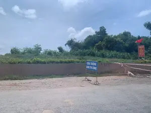 画像からのテキスト記録：。1. 青い看板：「工事中」。2. 赤いパネル：「ベトナム共産党万歳。」。3. コンクリートの壁に手書きの数字「9.13」があります。風景と活動の説明：。写真は道端の風景を撮影したもので、工事中の現場のようです。- 場所：道端のエリアで、農村部または郊外の可能性があります。手前はアスファルトの道路です。- 活動：このエリアは工事中の現場と特定され、赤と白の警告テープで一部が囲われています。人や建設活動は見られません。- 写真の中の物体：青い工事現場の看板、上部に赤い旗が付いた赤い政治宣伝パネル、低いコンクリートの壁、そして背後に生い茂る多くの木々や雑草。空は青く澄んでおり、雲があります