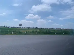 Bức ảnh chụp một khung cảnh ở vùng nông thôn ven đường. Ở tiền cảnh là mặt đường nhựa. Dọc theo lề đường có các cọc tiêu giao thông sơn màu trắng và đỏ. Có một biển báo được dựng lên, trên đó ghi rõ dòng chữ: 'KHU VỰC CẤM HỌP CHỢ'. Phía sau biển báo là một cánh đồng nông nghiệp rộng lớn. Ở phía xa là một khu dân cư hoặc một ngôi làng với nhiều ngôi nhà. Bầu trời trong xanh và có nhiều mây trắng.。（日本語訳）。写真は、道端の田舎の風景を写しています。手前にはアスファルトの路面があります。路肩に沿って、白と赤に塗られた視線誘導標があります。標識が一つ立てられており、そこには「市場開催禁止区域」という文字がはっきりと書かれています。標識の後ろには、広大な農地が広がっています。遠くには、多くの家々がある住宅地または村があります。空は青く澄んでおり、白い雲が多く浮かんでいます。