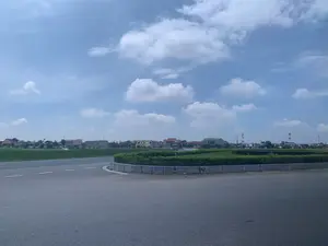 Khung cảnh chụp một vòng xuyến giao thông trên một con đường nhựa ở khu vực ngoại ô hoặc nông thôn. Vòng xuyến được xây bằng bê tông và trồng cây bụi xanh tươi. Bên trái là một cánh đồng lúa xanh mướt. Ở phía xa, có thể thấy nhiều ngôi nhà dân với các kiểu mái khác nhau, xen kẽ với cây cối. Một chiếc xe tải màu trắng đang di chuyển trên con đường phía xa. Có một số biển quảng cáo và một cấu trúc giống như cổng chào nhưng không thể đọc rõ chữ viết. Bầu trời trong xanh, có nhiều mây trắng.。（日本語訳）。郊外または田舎のアスファルト道路にある交通ラウンドアバウトを撮影した風景。ラウンドアバウトはコンクリートで造られ、青々とした低木が植えられている。左側には青々とした水田が広がっている。遠くの道路を白いトラックが一台走行している。いくつかの広告看板と、歓迎ゲートのような構造物があるが、書かれている文字ははっきりと読み取れない。空は青く澄み渡り、白い雲が多く浮かんでいる。