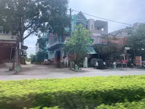 Bức ảnh chụp một con đường tại một khu dân cư ở Việt Nam, với nhiều ngôi nhà ống và cửa hàng san sát nhau. Tiền cảnh là một hàng rào cây xanh bị mờ, cho thấy ảnh được chụp từ một phương tiện đang di chuyển. Có nhiều cây xanh và dây điện dọc theo con đường. Nhiều xe máy đang đậu trước các cửa hàng, và một chiếc ô tô đang đậu trong một con hẻm nhỏ. Một vài người đang đứng gần các cửa tiệm. Các biển hiệu có thể đọc được bao gồm: 'CÚP 50' trên một biển hiệu màu đỏ, có thể là tiệm sửa xe. Một biển hiệu màu xanh lá cây ghi 'SỬA XE CHUYÊN...'. Một biển hiệu đứng màu trắng có chữ 'MAY' và 'VÉ SỐ'. Một biển hiệu màu cam bị che khuất một phần có chữ '... GIA DỤNG - HÀNG ... CHO TRẺ EM', gợi ý một cửa hàng bán đồ gia dụng và đồ cho trẻ em. Một biển hiệu khác ở phía xa ghi 'HÙNG TOÀN'.。（日本語訳）。写真はベトナムのある住宅街の通りを写したもので、多くのチューブハウス（縦長の家）や商店が隣接して建ち並んでいます。前景にはぼやけた緑の生垣があり、写真が移動中の乗り物から撮影されたことを示しています。通りに沿って多くの緑の木々と電線があります。多くのバイクが店の前に停められており、一台の自動車が小さな路地に停まっています。数人の人々が店の近くに立っています。読み取れる看板には、赤い看板に「CÚP 50」と書かれたものがあり、これはバイク修理店かもしれません。緑色の看板には「SỬA XE CHUYÊN...」（バイク修理専門...）と書かれています。白い立て看板には「MAY」（縫製）と「VÉ SỐ」（宝くじ）の文字があります。部分的に隠れているオレンジ色の看板には「... GIA DỤNG - HÀNG ... CHO TRẺ EM」（...家庭用品 - 子供向け...商品）という文字があり、家庭用品と子供用品を販売する店であることを示唆しています。遠くにある別の看板には「HÙNG TOÀN」と書かれています。