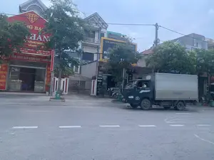 Bối cảnh là một con đường phố tại một khu vực đô thị ở Việt Nam, có thể là ở Thanh Hóa dựa trên thông tin trên biển hiệu. Một chiếc xe tải thùng màu xanh đậm, nhãn hiệu THACO, đang di chuyển trên đường, do một người đàn ông đội nón lá điều khiển. Hai bên đường là các tòa nhà và cửa hàng kinh doanh. Các văn bản có thể nhận dạng được bao gồm:。1. Biển hiệu lớn màu đỏ của một tiệm vàng: 'VÀNG BẠC HẢI THÁI', 'TRUNG TÂM MUA BÁN VÀNG BẠC TRANG SỨC', địa chỉ '145 - KCN ĐỊNH LIÊN - YÊN ĐỊNH - THANH HÓA', và 'DỊCH VỤ CHUYỂN TIỀN NHANH 24/7'.。2. Biển hiệu cửa hàng sửa chữa: 'SỬA CHỮA CHUYÊN NGHIỆP'.。3. Biển hiệu cửa hàng tạp hóa: 'TẠP HÓA TRUNG THỦY'.。4. Biển hiệu màu vàng: 'CỬA HÀNG ĐIỆN...' (chữ bị che khuất).。Dọc vỉa hè có cây xanh, một chiếc xe máy đang đậu và một thùng rác công cộng màu xanh lá cây.。（日本語訳）。背景はベトナムの都市部にある街路で、看板の情報に基づくとタインホア省である可能性があります。濃い青色のタコ（THACO）ブランドの箱型トラックが道路を走行しており、ノンラーをかぶった男性が運転しています。道路の両側には建物や商店が並んでいます。識別可能なテキストには以下が含まれます：。1. ある金店の大きな赤い看板：「VÀNG BẠC HẢI THÁI」、「TRUNG TÂM MUA BÁN VÀNG BẠC TRANG SỨC」、住所「145 - KCN ĐỊNH LIÊN - YÊN ĐỊNH - THANH HÓA」、そして「DỊCH VỤ CHUYỂN TIỀN NHANH 24/7」。2. 修理店の看板：「SỬA CHỮA CHUYÊN NGHIỆP」。3. 雑貨店の看板：「TẠP HÓA TRUNG THỦY」。4. 黄色い看板：「CỬA HÀNG ĐIỆN...」（文字が隠れている）。歩道沿いには緑の木々、駐車しているバイク1台、そして緑色の公共ゴミ箱があります。