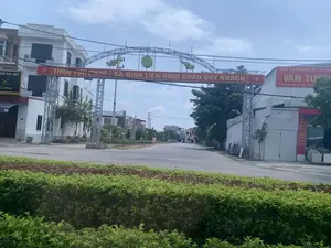 Bối cảnh là một con đường trải nhựa tại lối vào một khu dân cư ở Việt Nam, có thể là một thôn hoặc xã. Điểm nổi bật chính là một cổng chào lớn bằng kết cấu thép bắc ngang qua đường. Trên cổng có một biểu ngữ màu đỏ với dòng chữ vàng: 'THÔN VỰC PHÁC - XÃ ĐỊNH LIÊN KÍNH CHÀO QUÝ KHÁCH'. Trên cổng chào còn có các biểu tượng trang trí như búa liềm, ngôi sao vàng, hình con rồng và trống đồng. Hai bên đường là các tòa nhà nhiều tầng. Bên phải là một cửa hàng kinh doanh vật liệu xây dựng với biển hiệu 'TRUNG TÂM PHÂN PHỐI VẬT LIỆU XÂY DỰNG VĂN TÚ', địa chỉ 'ĐC: KCN ĐỊNH LIÊN - YÊN ĐỊNH'. Một biển hiệu dọc màu đỏ cũng liệt kê các mặt hàng như 'MÀNG SẮT THÉP SƠN & CHỐNG THẤM CAO CẤP'. Bên trái là một tòa nhà khác có biển hiệu 'ĐỒ ĂN VẶT Coffee' và số điện thoại. Con đường vắng vẻ, không có người và phương tiện qua lại. Cảnh quan xung quanh bao gồm cây xanh và nhiều đường dây điện.。（日本語訳）。背景はベトナムのある住宅地の入り口にあるアスファルト舗装された道で、村か集落である可能性があります。主な特徴は、道をまたぐ鉄骨造りの大きな歓迎ゲートです。ゲートの上には、「ヴック・ファック村 - ディン・リエン社 お客様を歓迎いたします」と黄色い文字で書かれた赤い横断幕があります。歓迎ゲートには、鎌と槌、金色の星、龍の絵、銅鼓といった装飾的なシンボルもあります。道の両側には複数階建ての建物があります。右側には、「ヴァン・トゥー建築資材流通センター」、住所「住所：ディン・リエン工業団地 - イエン・ディン」という看板を掲げた建築資材店があります。赤い縦長の看板には、「高級鉄鋼塗装＆防水材」といった商品も記載されています。左側には、「軽食・コーヒー」という看板と電話番号が掲げられた別の建物があります。道は閑散としており、人や乗り物の往来はありません。周囲の景観には、緑樹と多くの電線が含まれています。