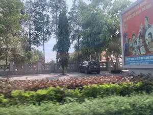 Bức ảnh chụp cảnh ven đường từ một phương tiện đang di chuyển. Nổi bật là một tấm biển cổ động lớn màu đỏ có nội dung: Phần trên là khẩu hiệu 'THI ĐUA LÀ YÊU NƯỚC, YÊU NƯỚC THÌ PHẢI THI ĐUA'. Phần dưới màu xanh có thông tin của một công ty, đọc được một phần là 'CÔNG TY TRUYỀN THÔNG & DU LỊCH...' và số điện thoại 'HOTLINE: 0977.75.1111 - 0917.75.1111'. Phía sau tấm biển là một khoảng sân có hàng rào bê tông kiên cố, nơi một chiếc ô tô SUV màu đen đang đỗ. Xung quanh có nhiều cây xanh cao lớn. Hàng cây bụi ở tiền cảnh bị mờ nhòe do hiệu ứng chuyển động.。（日本語訳）。移動中の乗り物から撮影された道端の風景写真です。目立っているのは、以下の内容が書かれた大きな赤いスローガン看板です。上部は「競争は愛国であり、愛国であるなら競争しなければならない」というスローガンです。下部の青い部分には会社の情報があり、一部「CÔNG TY TRUYỀN THÔNG & DU LỊCH...」と電話番号「HOTLINE: 0977.75.1111 - 0917.75.1111」が読み取れます。看板の後ろには頑丈なコンクリートの塀がある庭があり、そこには黒いSUV車が1台駐車しています。周囲には背の高い緑の木々がたくさんあります。前景の低木の列は、モーションエフェクトによりぼやけています。