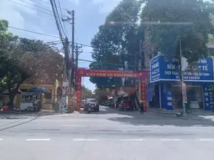 Bức ảnh chụp một con đường nhựa vào ban ngày tại một khu dân cư ở Việt Nam. Bầu trời có mây và nhiều cây xanh ở hai bên đường. Nổi bật nhất là một chiếc cổng chào màu đỏ có biểu tượng búa liềm và ngôi sao, bắc ngang qua đường với dòng chữ lớn: 'KHU DÂN CƯ KHU PHỐ 5'. Phía trên là dòng chữ nhỏ hơn 'ỦY BAN NHÂN DÂN THỊ TRẤN...'.。Hai bên đường là các tòa nhà cao tầng san sát. Bên phải là một cửa hàng lớn có mặt tiền màu xanh dương. Biển hiệu của cửa hàng ghi 'QUẤY THUỐC HẢI HẢI', chuyên bán 'BỈM - SỮA', 'THỜI TRANG TRẺ EM - ĐỒ SƠ SINH'. Các biển hiệu khác cũng cho thấy cửa hàng bán các sản phẩm 'Y TẾ CHÍNH HÃNG' và có ghi số điện thoại 'ĐT: 0368 334 333'. Một người phụ nữ đội nón lá đang đi bộ trên vỉa hè phía trước cửa hàng này.。Bên trái đường, dưới bóng cây, có một xe đẩy bán đồ ăn rong, trên xe có chữ 'XÔI'. Một người đang ngồi cạnh xe đẩy. Trên đường có một vài chiếc ô tô và xe máy đang đỗ. Hệ thống dây điện chằng chịt trên các cột điện là một hình ảnh đặc trưng của đường phố Việt Nam.。（日本語訳）。この写真は、ベトナムのある住宅街の日中のアスファルト道路を写したものです。空は曇っており、道の両側には多くの緑の木々があります。最も目立つのは、鎌と槌と星のシンボルがある赤い歓迎ゲートで、道をまたいでおり、「5区住宅街」という大きな文字が書かれています。その上には、より小さな文字で「…町人民委員会」と書かれています。道の両側には高層ビルが密集しています。右側には、青い正面をした大きな店があります。店の看板には「ハイハイ薬局」と書かれており、「おむつ・ミルク」、「子供服・ベビー用品」を専門に販売しています。他の看板にも、この店が「正規品医療」製品を販売していること、そして電話番号「電話：0368 334 333」が記載されていることが示されています。この店の前の歩道を、ノンラーをかぶった一人の女性が歩いています。道の左側、木陰に、屋台のカートがあり、そのカートには「ソイ」という文字が書かれています。一人の人がカートの隣に座っています。路上には数台の自動車とバイクが駐車しています。電柱の上で複雑に絡み合った電線は、ベトナムの街路の特特徴的な光景です。