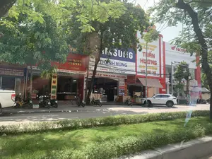 Bức ảnh chụp một con phố thương mại vào ban ngày, có nhiều cây xanh lớn che bóng mát. Dọc theo con đường là một dãy các cửa hàng kinh doanh san sát nhau.。Các thông tin văn bản có thể đọc được trên các biển hiệu bao gồm:。- Cửa hàng "ĐIỆN MÁY THANH HÙNG" với logo "SAMSUNG" lớn. Biển hiệu ghi rõ địa chỉ "...QUÁN LÀO, YÊN ĐỊNH, THANH HÓA" và số điện thoại "ĐT: 0912.384.269". Cửa hàng chuyên "TƯ VẤN LẮP ĐẶT: - BỘ DÀN KARAOKE - ÂM THANH LOA SÂN KHẤU - HỘI TRƯỜNG - ĐIỀU HÒA - MÁY GIẶT - TIVI - TỦ LẠNH - ĐIỆN GIA DỤNG". Các thương hiệu khác như Panasonic, Hitachi, Funiki, Sony, LG cũng được quảng cáo.。- "DOANH NGHIỆP VÀNG BẠC TUẤN HƯƠNG" kinh doanh vàng "14K - 18K - 24K", có địa chỉ tại "KHU 5 TT QUÁN LÀO, YÊN ĐỊNH, TH...".。- Tiệm "VÀNG BẠC ĐÁ QUÝ HOÀNG ANH" với biển hiệu màu đỏ, cam kết "Chất lượng - Bảo hành". Một biển hiệu dọc ghi "VÀNG BẠC HOÀNG ANH 9999".。- Một đại lý xe máy "HONDA" (HEAD) của "CÔNG TY TNHH...".。- Một cửa hàng ở phía bên trái có biển hiệu "CHÚNG TÔI GIÚP MẸ CHĂM SÓC CHO BÉ".。Về cảnh vật và hoạt động, đây là một khu phố kinh doanh. Có nhiều xe máy đang đậu trên vỉa hè trước các cửa hàng. Một chiếc ô tô SUV màu trắng đang đỗ bên lề đường. Phía bên trái có một phần của chiếc xe bán tải màu trắng. Con đường có dải phân cách ở giữa được trồng cỏ và cây bụi. Khung cảnh thể hiện một hoạt động kinh doanh thường nhật trên một con phố tại một thị trấn ở Việt Nam.。（日本語訳）。日中に撮影された商店街の写真で、大きな緑の木々が日陰を作っています。道沿いには、商店がずらりと軒を連ねています。看板から読み取れる文字情報は以下の通りです：。- 大きな「SAMSUNG」のロゴがある「ĐIỆN MÁY THANH HÙNG」店。看板には住所「...QUÁN LÀO, YÊN ĐỊNH, THANH HÓA」と電話番号「ĐT: 0912.384.269」が明記されています。この店は「コンサルティング・設置： - カラオケセット - ステージスピーカー音響 - ホール - エアコン - 洗濯機 - テレビ - 冷蔵庫 - 家庭用電化製品」を専門としています。パナソニック、日立、フニキ、ソニー、LGなどの他のブランドも宣伝されています。- 「DOANH NGHIỆP VÀNG BẠC TUẤN HƯƠNG」は「14K - 18K - 24K」の金を扱っており、住所は「KHU 5 TT QUÁN LÀO, YÊN ĐỊNH, TH...」です。- 赤い看板の「VÀNG BẠC ĐÁ QUÝ HOÀNG ANH」店は、「品質 - 保証」を約束しています。縦長の看板には「VÀNG BẠC HOÀNG ANH 9999」と書かれています。- 「CÔNG TY TNHH...」の「HONDA」バイク代理店（HEAD）。- 左側にある店には「お母さんの赤ちゃんケアをお手伝いします」という看板があります。風景と活動については、ここは商店街です。店の前の歩道には多くのバイクが停まっています。白いSUV車が路肩に駐車しています。左側には白いピックアップトラックの一部が見えます。道路の中央には草や低木が植えられた中央分離帯があります。この光景は、ベトナムのある町の通りでの日常的な商業活動を表しています。