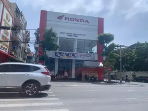 Bức ảnh chụp một con phố vào ban ngày, tập trung vào một tòa nhà lớn là đại lý xe máy Honda. Trên mặt tiền tòa nhà có logo 'HONDA' màu đỏ nổi bật, bên dưới là tên công ty 'CÔNG TY TNHH TM XUÂN THÀNH PHÁT' và địa chỉ 'THIỆU YÊN 1'. Phía trước cửa hàng có treo một băng rôn quảng cáo với khẩu hiệu 'RƯỚC QUÀ ĐÚNG GU - Phượt hè đứng đỉnh' cho chương trình diễn ra từ 01/07/2024 đến 30/09/2024. Một quầy trưng bày màu đỏ phía trước ghi 'HỆ THỐNG HEAD THIÊU YÊN', 'HEAD 21032 - THIỆU YÊN 1' và 'TRƯNG BÀY VÀ GIỚI THIỆU SẢN PHẨM MỚI'. Cảnh vật cho thấy hoạt động kinh doanh đang diễn ra. Có nhiều xe máy được trưng bày bên trong và đậu bên ngoài. Một chiếc ô tô SUV màu trắng đang ở trên đường, phía trước có vạch kẻ đường cho người đi bộ. Vài người dân đang ở trên vỉa hè. Một tấm biển nhỏ gần đó đăng tin 'TUYỂN NHÂN VIÊN BÁN HÀNG' kèm số điện thoại 'LH: 0889 00 9999'. Tòa nhà bên cạnh có biển hiệu 'DOANH NGHIỆP VÀNG TUẤN HƯƠNG'.。（日本語訳）。日中の通りを撮影した写真で、ホンダのバイク代理店である大きな建物に焦点が当てられています。建物の正面には目立つ赤い「HONDA」のロゴがあり、その下には会社名「CÔNG TY TNHH TM XUÂN THÀNH PHÁT」と住所「THIỆU YÊN 1」があります。店の前には、2024年7月1日から2024年9月30日まで開催されるプログラムの「好みに合ったプレゼントをゲット - 最高の夏のツーリング」というスローガンが書かれた広告バナーが掲げられています。前面にある赤い展示カウンターには、「HỆ THỐNG HEAD THIÊU YÊN」、「HEAD 21032 - THIỆU YÊN 1」、そして「新製品の展示と紹介」と書かれています。風景は、営業活動が行われていることを示しています。店内には多くのバイクが展示され、外にも駐車されています。道路には白いSUV車が1台あり、その前には横断歩道の白線があります。歩道には数人の人がいます。近くの小さな看板には、「販売スタッフ募集」という告知と電話番号「連絡先: 0889 00 9999」が記載されています。隣の建物には「DOANH NGHIỆP VÀNG TUẤN HƯƠNG」という看板があります。