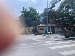 Hình ảnh ghi lại một góc phố vào một ngày nắng, có vạch kẻ đường dành cho người đi bộ. Bên phải đường là một chiếc máy xúc lớn màu đen mang nhãn hiệu "VOLVO". Phía sau là một tòa nhà có các biển hiệu "AGRIBANK", "AGRIBANK PLUS", và "AUTOBANK", cùng với một áp phích quảng cáo ghi "Dễ dùng Dễ dàng". Tại góc đường có một chốt gác nhỏ, bên cạnh là một biển báo đường phố màu xanh ghi "ĐƯƠNG TRIỀN QUỐC BÁT". Một cột mốc xi măng ghi "DƯƠNG", "Km 0", và "SÉT 5 Km". Một người đàn ông đang ngồi trên cột mốc, trong khi vài người khác ngồi gần chốt gác. Khung cảnh có cây xanh và cột điện. Một phần của hình ảnh ở phía trước bên trái bị mờ.。（日本語訳）。晴れた日の街角を写した写真で、歩行者用の横断歩道があります。道の右側には、「VOLVO」ブランドの大きな黒いショベルカーがあります。その後ろには、「AGRIBANK」、「AGRIBANK PLUS」、「AUTOBANK」という看板がある建物と、「使いやすい 簡単」と書かれた広告ポスターがあります。通りの角には小さな警備員の詰め所があり、その隣には「ĐƯƠNG TRIỀN QUỐC BÁT」と書かれた青い道路標識があります。セメント製のキロポストには、「DƯƠNG」、「Km 0」、「SÉT 5 Km」と書かれています。一人の男性がキロポストの上に座っており、他の数人が警備員の詰め所の近くに座っています。風景には緑の木々と電柱があります。写真の左前の一部がぼやけています。