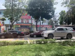 背景は都市部のアスファルト道路で、多くの交通車両があります。写真は移動中の乗り物から撮影されたものです。写真の中で目立っているのは、アグリバンク銀行の青い建物です。記録されている文字情報は以下の通りです：。- 銀行の看板には、「AGRIBANK」、「NGÂN HÀNG NÔNG NGHIỆP & PTNT VIỆT NAM」（ベトナム農業農村開発銀行）、「VIETNAM BANK FOR AGRICULTURE AND RURAL DEVELOPMENT」、そして「CHI NHÁNH HUYỆN YÊN ĐỊNH THANH HÓA」（タインホア省イエンディン県支店）と書かれています。- 黄色い文字の赤い横断幕には、「ĐẢNG CỘNG SẢN VIỆT NAM QUANG VINH MUÔN NĂM。」（ベトナム共産党万歳。）と書かれています。- 広告バナーには、「CÀI Đ... THU THẬP SINH TRẮC HỌC trên BANK PLUS」（BANK PLUSで生体認証情報を収集するためにインストール...）と書かれています。- 左側には「ATM」と「AUTOBANK」の看板があります。風景と活動の描写：。- 場所：沿道に建物と緑樹がある街路。具体的には、タインホア省イエンディン県のアグリバンク銀行支店の前です。- 活動：道路交通。一台の銀色のSUV車が通り過ぎている一方で、一台の黒い車、一台の濃い赤色の車、そして一台のバイクが路肩に駐車しているか、ゆっくりと移動しています。- その他の物体：多くの大きな緑樹、白いフェンス、拡声器付きの電柱