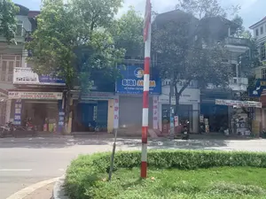 Bối cảnh là một con phố tại thị trấn Quán Lào, huyện Yên Định, tỉnh Thanh Hóa. Hai bên đường là những ngôi nhà ống nhiều tầng, tầng trệt được sử dụng làm mặt bằng kinh doanh. Có nhiều biển hiệu của các cửa hàng có thể đọc được:。- Biển hiệu lớn màu trắng của cửa hàng điện tử: "EZVIZ... LAPTOP - MÁY IN - CAMERA. Địa chỉ: 226 - Đường Mùng 10 Tháng 6 - TT. Quán Lào - Yên Định - Thanh Hóa. Điện thoại 0904771136 - 0915843011".。- Biển hiệu màu vàng của một đại lý nông nghiệp: "ĐẠI LÝ KỸ SƯ NÔNG NGHIỆP: NGUYỄN XUÂN PHƯƠNG - GIỐNG CÂY TRỒNG. LÚA - NGÔ - ĐẬU - LẠC - HẠT RAU - CÂY ĂN QUẢ (BẢO HÀNH CHẤT LƯỢNG)". Địa chỉ: "229 - TK 4 - TT QUÁN LÀO".。- Biển hiệu màu xanh của cửa hàng mẹ và bé "BIBI MOM - HỆ THỐNG CỬA HÀNG... BẦU" tại "SN 222 - KHU 4 - TT QUÁN LÀO - YÊN ĐỊNH - TH". Cửa hàng này có cửa cuốn đang đóng và có biển dọc ghi "TRẢ GÓP 0%".。- Một cửa hàng có biển hiệu "...A STORE" và biển dọc "SỬA CHỮA LẤY NGAY".。- Một tấm bạt của cửa hàng "Hạnh Nhi" chuyên "SỬA CHỮA - MUA BÁN ĐIỆN LẠNH - ĐIỆN DÂN DỤNG".。Hoạt động: Khung cảnh khá yên tĩnh, một số cửa hàng đóng cửa cuốn. Có vài chiếc xe máy đang đậu trước các cửa hàng. Vật thể: Các ngôi nhà, cửa hàng, xe máy, cây xanh, cột báo hiệu sọc đỏ trắng ở tiền cảnh, và các biển quảng cáo.。（日本語訳）。背景は、タインホア省イエンディン県クアンラオ町の通りです。道の両側には多層階のチューブハウスが並び、1階は店舗として使用されています。読み取れる店の看板がいくつかあります：。- 電子機器店の大きな白い看板：「EZVIZ... ノートパソコン - プリンター - カメラ。住所：タインホア省イエンディン県クアンラオ町6月10日通り226番地。電話番号 0904771136 - 0915843011」。- 農業代理店の黄色い看板：「農業技術者代理店：グエン・スアン・フオン - 作物の種子。稲 - トウモロコシ - 豆 - 落花生 - 野菜の種 - 果樹（品質保証）」。住所：「クアンラオ町4区229番地」。- 「SN 222 - KHU 4 - TT QUÁN LÀO - YÊN ĐỊNH - TH」にあるマタニティ・ベビー用品店「BIBI MOM - 店舗システム...マタニティ」の青い看板。この店はシャッターが閉まっており、「分割払い0%」と書かれた縦看板があります。- 「...A STORE」という看板と、「即日修理」という縦看板がある店。- 「Hạnh Nhi」店の、「冷凍空調設備 - 家庭用電化製品の修理・売買」を専門とするシート看板。活動：風景はかなり静かで、いくつかの店はシャッターが閉まっています。店の前に数台のバイクが停まっています。物体：家、店、バイク、緑の木々、前景にある赤と白の縞模様の標識柱、そして広告看板。