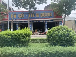 Bản ghi văn bản từ hình ảnh:。Trên biển hiệu lớn màu xanh dương với chữ màu vàng: ĐỒ GỖ - TIẾN DŨNG.。Bên dưới là biển hiệu: CỬA HÀNG ĐỒ GỖ TIẾN DŨNG.。Trên các biển hiệu dọc treo ở cột: ĐỒ GỖ SẢN XUẤT, BÀN THỜ, BÀN GHẾ, GIƯỜNG TỦ, CHĂN GA GỐI ĐỆM, THIẾT BỊ VĂN PHÒNG, KỆ GỖ.。Trên biển hiệu của tòa nhà bên trái: Hotline 0963 094 818.。Trên biển hiệu bên phải có thông tin địa chỉ và số điện thoại: ĐC: Khu 4 - TT. Quán Lào - Yên Định, ĐT: 0904 625 344 và một số điện thoại khác là 0975 675...。Mô tả cảnh quan và hoạt động:。Đây là hình ảnh chụp một cửa hàng đồ gỗ tên là "Tiến Dũng" nằm ở ven đường. Cửa hàng có mặt tiền rộng, với một biển hiệu lớn màu xanh và vàng nổi bật. Phía trước cửa hàng, một số sản phẩm nội thất bằng gỗ như bàn, ghế và kệ được trưng bày để bán. Theo thông tin trên biển hiệu, cửa hàng chuyên bán các mặt hàng như giường, tủ, bàn ghế, bàn thờ, chăn ga gối đệm và thiết bị văn phòng. Khung cảnh cho thấy đây là một khu vực kinh doanh. Phía trước có nhiều cây xanh và bụi rậm, cho thấy bức ảnh có thể được chụp từ bên kia đường hoặc từ một phương tiện đang di chuyển.。（日本語訳）。画像からの文字起こし：。黄色い文字の大きな青い看板に「木製家具 - TIEN DUNG」。その下には「TIEN DUNG木製家具店」という看板があります。柱に掛けられた縦長の看板には、「木製家具製造」「祭壇」「テーブル・椅子」「ベッド・タンス」「寝具（毛布・シーツ・枕・マットレス）」「オフィス用品」「木製棚」。左側の建物の看板に「ホットライン 0963 094 818」。右側の看板には住所と電話番号の情報があります。「住所：イエンディン県クアンラオ町第4地区、電話：0904 625 344」、そしてもう一つの電話番号は「0975 675...」。風景と状況の説明：。これは道端にある「ティエン・ズン」という名前の木製家具店を撮影した写真です。店は間口が広く、目立つ青と黄色の大きな看板があります。店の前には、テーブル、椅子、棚などの木製家具製品がいくつか販売のために展示されています。看板の情報によると、この店はベッド、タンス、テーブル・椅子、祭壇、寝具（毛布・シーツ・枕・マットレス）、オフィス用品などの商品を専門に販売しています。この光景は、ここが商業地域であることを示しています。前景には多くの緑の木々や茂みがあり、写真が道路の向こう側から、または移動中の乗り物から撮影された可能性を示唆しています。