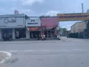 Đây là khung cảnh một con đường ở Việt Nam với một dãy cửa hàng kinh doanh san sát nhau. Bầu trời có mây. Các cửa hàng và biển hiệu có thể được xác định như sau:。1.  **Chữ viết được ghi lại:**。*   **Cửa hàng Orchid:** "Orchid", "QUẢN LÀO, YÊN ĐỊNH, THANH HÓA", và số điện thoại "033 501 3993".。*   **Cửa hàng MONALIZA:** Logo "M" và chữ "MONALIZA". Địa chỉ: "Đ/C: KP4, QUÁN LÀO, YÊN ĐỊNH, THANH HÓA". Số điện thoại: "0356 280 121". Có một băng rôn quảng cáo lớn màu đỏ ghi: "TƯNG BỪNG KHAI TRƯƠNG", "SALE OFF 50%", "Thời gian: Từ 24/03 - 01/04".。*   **Cửa hàng Thỏ:** "Thời Trang Trẻ Em", "Thỏ..." cùng logo hình con thỏ. Địa chỉ: "KHU 4 - TT. QUÁN LÀO". Số điện thoại: "0908 189 578". Có băng rôn "MỪNG LỄ 30/4" và "SALE OFF 10%".。*   **Biển hiệu Bến xe:** Một cổng chào lớn màu vàng ghi "CÔNG TY TNHH MINH QUANG", "BẾN XE KHÁCH QUÁN LÀO", "Kính Chào Quý Khách。". Địa chỉ: "Khu 4 - TT. Quán Lào - Yên Định - TH". Số điện thoại: "ĐT: 0948.166.289".。*   **Biển hiệu trên nóc nhà:** Có một biển hiệu màu đỏ ghi "MAI ANH" trên nóc tòa nhà của cửa hàng Orchid.。2.  **Mô tả cảnh vật và hoạt động:**。*   **Loại địa điểm:** Đây là một khu phố thương mại trên một con đường tại Thị trấn Quán Lào, huyện Yên Định, tỉnh Thanh Hóa. Bên phải có cổng vào của một bến xe khách.。*   **Hoạt động:** Các cửa hàng đang hoạt động kinh doanh. Cửa hàng MONALIZA đang trong thời gian khai trương với chương trình giảm giá. Cửa hàng Thỏ cũng có chương trình khuyến mãi cho dịp lễ 30/4. Có một vài người đang đứng trước cửa hàng thời trang trẻ em, có thể đang mua sắm hoặc xem hàng. Một số xe máy đang đỗ trên vỉa hè, cho thấy có khách hàng hoặc nhân viên tại các cửa hàng.。*   **Vật thể:** Có ba cửa hàng thời trang, trong đó hai cửa hàng bán đồ người lớn ("Orchid", "MONALIZA") và một cửa hàng bán đồ trẻ em ("Thỏ"). Nhiều ma-nơ-canh được trưng bày trong và ngoài cửa hàng để giới thiệu quần áo. Các băng rôn quảng cáo được treo để thu hút khách hàng. Một số xe máy (xe ga và xe số) đang đỗ. Phía xa có một cột thu phát sóng viễn thông.。（日本語訳）。これはベトナムのある通りの風景で、商店がずらりと並んでいます。空には雲があります。各店舗と看板は以下のように特定できます。1.  **記録された文字：**。*   **Orchid店：** 「Orchid」、「QUẢN LÀO, YÊN ĐỊNH, THANH HÓA」、および電話番号「033 501 3993」。*   **MONALIZA店：** 「M」のロゴと「MONALIZA」の文字。住所：「Đ/C: KP4, QUÁN LÀO, YÊN ĐỊNH, THANH HÓA」。電話番号：「0356 280 121」。大きな赤い広告バナーがあり、「グランドオープン」、「50%オフセール」、「期間：3月24日～4月1日」と書かれています。*   **Thỏ店：** 「子供服」、「Thỏ（ウサギ）...」とウサギの形のロゴ。住所：「KHU 4 - TT. QUÁN LÀO」。電話番号：「0908 189 578」。「4月30日祝日記念」と「10%オフセール」のバナーがあります。*   **バスターミナルの看板：** 大きな黄色のウェルカムゲートに「MINH QUANG有限会社」、「QUÁN LÀO旅客バスターミナル」、「お客様を歓迎します。」と書かれています。住所：「Khu 4 - TT. Quán Lào - Yên Định - TH」。電話番号：「ĐT: 0948.166.289」。*   **屋上の看板：** Orchid店の建物の屋上に「MAI ANH」と書かれた赤い看板があります。2.  **風景と活動の説明：**。*   **場所の種類：** これはタインホア省イエンディン県クアンラオ町の通りにある商店街です。右側には旅客バスターミナルの入口ゲートがあります。*   **活動：** 各店舗は営業中です。MONALIZA店は開店記念期間中で割引キャンペーンを行っています。Thỏ店も4月30日の祝日に向けたプロモーションプログラムを実施しています。子供服店の前に数人が立っており、買い物をしているか商品を見ている可能性があります。歩道には数台のバイクが停まっており、店舗に客や従業員がいることを示しています。*   **物体：** 3つのファッション店があり、そのうち2店は大人向け（「Orchid」、「MONALIZA」）、1店は子供向け（「Thỏ」）です。多くのマネキンが服を紹介するために店内外に展示されています。広告バナーが客を引きつけるために掲げられています。数台のバイク（スクーターとマニュアル車）が停まっています。遠くには通信用の電波塔があります。