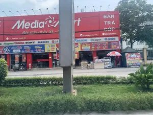 Bức ảnh chụp mặt tiền của siêu thị điện máy MediaMart tại Thị trấn Quán Lào, Thanh Hóa. Tòa nhà có màu đỏ đặc trưng với tên thương hiệu 'MediaMart' và dòng chữ 'điện máy - máy tính' được viết lớn. Nhiều biển hiệu quảng cáo của các hãng nổi tiếng như Kangaroo, TOSHIBA, SONY, SAMSUNG, LG, DAIKIN, Casper, GREE được trưng bày. Các áp phích quảng cáo các chương trình khuyến mãi như 'BÁN TRẢ GÓP', 'XẢ HÀNG GIÁ SỐC', 'đồng loạt giảm đến 50%', và 'mua điều hòa Tặng quà khủng đến 7 triệu' được treo khắp nơi. Giá của một số sản phẩm cũng được niêm yết công khai, ví dụ 'Tủ lạnh Inverter 185 Lít' giá 4.49 triệu đồng và 'Quạt cây' giá 299 nghìn đồng. Phía trước cửa hàng, có hai chiếc xe máy đang đỗ và nhiều thùng hàng điện tử được xếp chồng lên nhau. Một chiếc ô lớn màu đỏ trắng có logo MediaMart được dựng lên. Khung cảnh cho thấy một con đường với dải phân cách có cây xanh ở phía trước.。（日本語訳）。タインホア省クアンラオ町にある家電量販店メディアマートの正面を撮影した写真です。建物は特徴的な赤色で、「MediaMart」というブランド名と「家電 - パソコン」という文字が大きく書かれています。Kangaroo、TOSHIBA、SONY、SAMSUNG、LG、DAIKIN、Casper、GREEなどの有名メーカーの広告看板が多数展示されています。「分割払い販売」、「衝撃価格の在庫一掃セール」、「一斉に最大50%割引」、「エアコン購入で最大700万ドンの豪華プレゼント」といったプロモーションプログラムの広告ポスターが至る所に掲げられています。一部の商品の価格も公に表示されており、例えば「インバーター冷蔵庫185リットル」は449万ドン、「扇風機」は29万9千ドンです。店の前には、2台のバイクが停まっており、多くの電子機器の箱が積み重ねられています。MediaMartのロゴが入った赤と白の大きなパラソルが立てられています。風景は、手前に緑の木々がある中央分離帯のある道路を示しています。