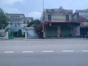 Bối cảnh là một con đường nhựa tại một khu đô thị, có thể là ở thị trấn Quán Lào, huyện Yên Định. Bên trái là tòa nhà trụ sở Bảo hiểm xã hội huyện Yên Định, với các biển hiệu và băng rôn tuyên truyền. Các dòng chữ có thể đọc được bao gồm: 'BẢO HIỂM XÃ HỘI HUYỆN YÊN ĐỊNH' và băng rôn 'HƯỚNG TỚI MỤC TIÊU THỰC HIỆN BẢO HIỂM XÃ HỘI, BẢO HIỂM Y TẾ TOÀN DÂN LÀ GÓP PHẦN XÂY DỰNG SỰ NGHIỆP AN SINH BỀN VỮNG。'. Bên phải là một dãy nhà phố thương mại. Một cửa hàng 'TEA & COFFEE' ở địa chỉ 'KHU 4 THỊ TRẤN QUÁN LÀO' đã đóng cửa cuốn và có treo biển 'CHO THUÊ NHÀ ĐT: 0945 363 818'. Tầng trên của tòa nhà này có biển hiệu 'Acoustic Oishi Live-Music'. Kế bên là văn phòng của 'CÔNG TY TNHH DYNAMIC' chuyên 'TƯ VẤN DU HỌC VÀ XUẤT KHẨU LAO ĐỘNG' tới Nhật Bản, Hàn Quốc, Úc, Đức, Canada, Pháp. Một vài chiếc xe máy đang đỗ trên vỉa hè và một người đang đi xe đạp trên đường. Cảnh vật yên tĩnh dưới bầu trời u ám.。（日本語訳）。背景は、都市部のアスファルト道路で、おそらくイエンディン県クアンラオ町にある。読み取れる文字には、「イエンディン県社会保険」と、「全国民の社会保険、医療保険の実施という目標に向かうことは、持続可能な社会保障事業の構築に貢献することである。」という横断幕が含まれる。右側には商店が並んでいる。この建物の上の階には「Acoustic Oishi Live-Music」という看板がある。その隣には、日本、韓国、オーストラリア、ドイツ、カナダ、フランスへの「留学コンサルティングと労働者派遣」を専門とする「DYNAMIC有限会社」のオフィスがある。数台のバイクが歩道に停まっており、一人が道路を自転車で走っている。曇り空の下、景色は静かである。