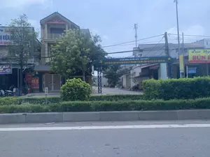 Bức ảnh chụp một con đường tại một khu dân cư ở Việt Nam, nổi bật là một cổng chào màu xanh lam dẫn vào một con ngõ. Trên cổng chào có các dòng chữ: 'TỔ AN NINH XÃ HỘI SỐ 8 - KHU PHỐ 4' và khẩu hiệu 'QUYẾT TÂM XÂY DỰNG KHU PHỐ GIÀU VỀ KINH TẾ, VỮNG MẠNH VỀ AN NINH CHÍNH TRỊ VÀ TRẬT TỰ AN TOÀN XÃ HỘI。'. Hai bên đường là những ngôi nhà nhiều tầng và các cửa hàng kinh doanh. Bên trái có một tiệm 'RỬA XE - Ô TÔ' cung cấp dịch vụ 'BỌT TUYẾT', 'VÁ SĂM, THAY DẦU' và một cửa hàng khác có biển hiệu '...Ụ KIỆN', 'IN DECAL'. Một vài chiếc xe máy đang đỗ gần đó. Bên phải là một quán ăn có biển hiệu màu vàng ghi 'Lực Ng... BÚN...' với địa chỉ 'Đ/c: Đường Mùng 10 Tháng 6 - Khu 4 - TT.Quán...'. Một biển báo đường phố màu xanh cũng xác nhận tên đường là 'ĐƯỜNG MÙNG 10 THÁNG 6'. Con đường chính có dải phân cách trồng cây xanh. Ở phía xa, dưới cổng chào, có một vài người đang đi bộ. Toàn cảnh thể hiện một khung cảnh sinh hoạt đời thường vào ban ngày.。（日本語訳）。ベトナムのある住宅街の道を撮影した写真で、路地へと続く青い歓迎ゲートが目立っている。歓迎ゲートには「第8社会治安班 - 第4街区」という文字と、「経済的に豊かな街区、政治的治安と社会安全秩序において強固な街区の建設を決意する。」というスローガンが書かれている。道の両側には多層階の家々や商店が並んでいる。左側には「洗車 - 自動車」店があり、「スノーフォーム」、「パンク修理、オイル交換」のサービスを提供しており、別の店には「...部品」、「デカール印刷」という看板がある。近くに数台のバイクが停まっている。右側には「Lực Ng... BÚN...」と書かれた黄色い看板の食堂があり、住所は「住所：6月10日通り - 第4区 - Quán...町」である。青い道路標識も通りの名前が「6月10日通り」であることを示している。主要道路には緑の木々が植えられた中央分離帯がある。遠くの歓迎ゲートの下では、数人が歩いている。全景は日中の日常的な生活風景を表している。