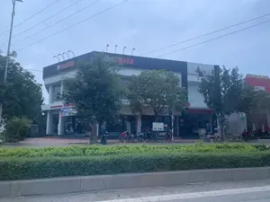 Bức ảnh chụp một đại lý xe máy lớn, hiện đại nằm ven đường. Đây là một cửa hàng của Yamaha, với biển hiệu lớn "YAMAHA" và "Yamaha Town" trên mặt tiền tòa nhà hai tầng. Phía trước cửa hàng có nhiều xe máy đang đậu và cây xanh. Qua cửa kính, có thể thấy xe máy được trưng bày bên trong.。Các dòng chữ có thể đọc được bao gồm:。- Trên tòa nhà chính: "YAMAHA", "Yamaha Town".。- Biển hiệu đứng: "ĐIỂM BÁN BẢO HIỂM Ô TÔ - XE MÁY".。- Trên tường: "TRUNG TÂM BẢO HÀNH VÀ DỊCH VỤ".。- Poster bên trong: "BOTH LIVES MAXIMIZED".。- Tòa nhà bên cạnh: "SYM", "SIÊU THỊ XE MÁY".。Khung cảnh cho thấy một hoạt động thương mại diễn ra bên đường. Có một dải phân cách với cây bụi ở tiền cảnh. Ở phía bên phải, một người đàn ông đang ngồi trên băng ghế. Bầu trời có vẻ u ám. Bên trái có một cột điện gắn loa phát thanh.。（日本語訳）。写真は、道端にある大きくて近代的なバイク代理店を写しています。これはヤマハの店舗で、2階建ての建物の正面には「YAMAHA」と「Yamaha Town」の大きな看板があります。店の前には多くのバイクが停められており、緑の木々があります。ガラスのドア越しに、中に展示されているバイクが見えます。読み取れる文字は以下の通りです：。- メインの建物：「YAMAHA」、「Yamaha Town」。- 立て看板：「自動車・バイク保険販売店」。- 壁面：「保証・サービスセンター」。- 中のポスター：「BOTH LIVES MAXIMIZED」。- 隣の建物：「SYM」、「バイクスーパーマーケット」。この光景は、道端で行われている商業活動を示しています。前景には低木のある中央分離帯があります。右側では、男性がベンチに座っています。空は曇っているようです。左側には放送用スピーカーが取り付けられた電柱があります。