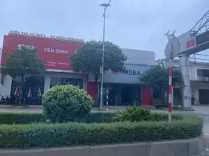 Bức ảnh chụp khung cảnh phía trước một tòa nhà thương mại, có vẻ là một siêu thị xe máy và xe điện. Trên tấm biển hiệu lớn màu đỏ có dòng chữ màu vàng: 'SIÊU THỊ XE MÁY - XE ĐIỆN YÊN ĐỊNH'. Bên dưới là các dòng chữ 'YÊN ĐỊNH', 'YÊN ĐỊNH - THANH HÓA' và số điện thoại '373 54 9999'. Bên cạnh, một phần khác của tòa nhà có biển hiệu của thương hiệu 'YADEA'. Nhiều xe máy và xe điện được trưng bày bên trong và phía trước cửa hàng. Phía trước có cây xanh và các bụi cây được cắt tỉa. Có một cột đèn đường năng lượng mặt trời. Ở phía bên phải, có một cấu trúc giống cổng chào với biểu ngữ màu đỏ có hình búa liềm và chữ 'ĐẢNG M...' nhưng không thể đọc hết. Cảnh vật cho thấy một con đường tại một khu vực đô thị hoặc thị trấn dưới bầu trời u ám.。（日本語訳）。商業ビルの正面の風景を写した写真で、バイクと電動バイクのスーパーマーケットのようです。大きな赤い看板には黄色い文字で「SIÊU THỊ XE MÁY - XE ĐIỆN YÊN ĐỊNH」と書かれています。その下には「YÊN ĐỊNH」、「YÊN ĐỊNH - THANH HÓA」、そして電話番号「373 54 9999」の文字があります。隣の建物の別の部分には、「YADEA」ブランドの看板があります。店内と店の前には多くのバイクと電動バイクが展示されています。正面には緑の木々と刈り込まれた低木があります。太陽光発電の街灯が1本あります。右側には、歓迎ゲートのような構造物があり、そこには鎌と槌の絵と「ĐẢNG M...」という文字が書かれた赤い横断幕がありますが、全文は読めません。風景は、曇り空の下、都市部または町の一本の道を示しています。