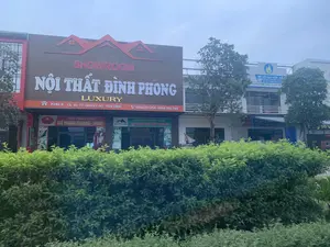 Bức ảnh chụp một dãy nhà phố thương mại với nhiều cửa hàng san sát nhau. Nổi bật nhất là một showroom lớn có biển hiệu màu nâu với dòng chữ "SHOWROOM NỘI THẤT ĐÌNH PHONG LUXURY". Trên biển hiệu có ghi địa chỉ "KHU 4, QL 45 TT QUÁN LÀO, YÊN ĐỊNH" và số điện thoại "0984201254 - 0968706789". Bên dưới, có hai biển hiệu nhỏ hơn cho thấy đây cũng là "NHÀ PHÂN PHỐI GHẾ MASAGE FUJIKIMA - TOYOKY" và "NHÀ PHÂN PHỐI ĐỘC QUYỀN NỆM CAO SU THIÊN NHIÊN TRUNG SƠN".。Bên phải là một cửa hàng khác với biển hiệu màu xanh dương, kinh doanh "ĐỒ GỖ CAO CẤP". Các dòng chữ khác trên biển hiệu bao gồm "NHẬN ĐẶT HÀNG VÀ THI CÔNG CÁC LOẠI BÀN GHẾ, TỦ, GIƯỜNG, LAN CAN, KỆ,..." và "...CÔNG NGHỆ XANH - TỰ HÀO THƯ...".。Khung cảnh cho thấy một con đường kinh doanh. Phía trước các tòa nhà là một hàng rào cây xanh. Bầu trời u ám. Không có người hoặc hoạt động cụ thể nào được ghi lại trong ảnh.。（日本語訳）。写真は、多くの店が隣接する商店街の一角を写しています。最も目立つのは、「SHOWROOM NỘI THẤT ĐÌNH PHONG LUXURY」という文字が書かれた茶色の看板がある大きなショールームです。看板には住所「KHU 4, QL 45 TT QUÁN LÀO, YÊN ĐỊNH」と電話番号「0984201254 - 0968706789」が記載されています。その下には、ここが「NHÀ PHÂN PHỐI GHẾ MASAGE FUJIKIMA - TOYOKY」と「NHÀ PHÂN PHỐI ĐỘC QUYỀN NỆM CAO SU THIÊN NHIÊN TRUNG SƠN」でもあることを示す、2つの小さな看板があります。右側には、「ĐỒ GỖ CAO CẤP」を商う、青い看板の別の店があります。看板の他の文言には、「NHẬN ĐẶT HÀNG VÀ THI CÔNG CÁC LOẠI BÀN GHẾ, TỦ, GIƯỜNG, LAN CAN, KỆ,...」および「...CÔNG NGHỆ XANH - TỰ HÀO THƯ...」が含まれています。この光景は、商業道路であることを示しています。建物の前には生け垣があります。空は曇っています。写真には人や特定の活動は写っていません。