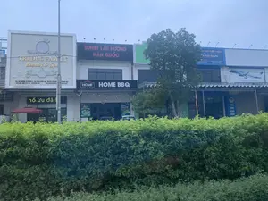 Bức ảnh chụp một dãy các cửa hàng và cơ sở kinh doanh liền kề nhau trên một con phố. Phía trước có một hàng rào cây xanh. Các biển hiệu có thể đọc được bao gồm:。1. Một cơ sở làm đẹp có tên "TRÍ ĐỨC FAMILY Beauty & Spa". Biển hiệu ghi rõ địa chỉ: "ĐỊA CHỈ: ĐƯỜNG 10 THÁNG 6 - THỊ TRẤN QUÁN LÀO - HUYỆN YÊN ĐỊNH - THANH HÓA", và thông tin liên hệ "HOTLINE: 0985 210 182 - 036 8282 969", "FB.COM/TRIDUCFA". Một bảng đèn LED chạy chữ "nổ ưu đãi。".。2. Một nhà hàng có tên "HOME BBQ" với biển hiệu lớn phía trên ghi "BUFFET LẨU NƯỚNG HÀN QUỐC".。3. Một cửa hàng có biển hiệu "IKO SAN" và "CHĂN - GA - GỐI - ĐỆM".。4. Một cửa hàng có biển quảng cáo của "Vietnam Airlines" và một biển hiệu nhỏ hơn ghi "YẾN SÀO KHÔ NHA".。Khung cảnh là một khu phố thương mại với các tòa nhà san sát. Có một cây xanh lớn, một cột điện, và một chiếc ô màu đỏ. Một vài chiếc xe ô tô đậu phía sau hàng rào cây.。（日本語訳）。写真は、ある通りに隣接して並ぶ一連の店舗や商業施設を撮影したものです。手前には緑の植え込みの列があります。読み取れる看板には以下のものが含まれます：。1. 「TRÍ ĐỨC FAMILY Beauty & Spa」という名前の美容施設。看板には住所：「ĐỊA CHỈ: ĐƯỜNG 10 THÁNG 6 - THỊ TRẤN QUÁN LÀO - HUYỆN YÊN ĐỊNH - THANH HÓA」（住所：6月10日通り - クアンラオ町 - イエンディン県 - タインホア省）、および連絡先情報「HOTLINE: 0985 210 182 - 036 8282 969」、「FB.COM/TRIDUCFA」が明記されています。「nổ ưu đãi。」（特典爆発。）という文字が流れるLED電光掲示板があります。2. 「HOME BBQ」という名前のレストランで、上部にある大きな看板には「BUFFET LẨU NƯỚNG HÀN QUỐC」（韓国式焼肉鍋ビュッフェ）と書かれています。3. 「IKO SAN」と「CHĂN - GA - GỐI - ĐỆM」（掛け布団 - シーツ - 枕 - マットレス）という看板がある店。4. 「Vietnam Airlines」の広告看板と、「YẾN SÀO KHÔ NHA」（乾燥燕の巣）と書かれた小さな看板がある店。風景は、建物が密集した商店街です。大きな緑の木が1本、電柱が1本、そして赤い傘が1つあります。植え込みの列の後ろに数台の自動車が駐車しています。