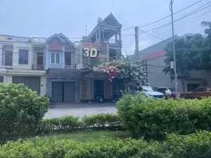 Bản ghi chữ viết: Trên tòa nhà ở trung tâm có một biển hiệu lớn màu vàng ghi "3D.com". Bên dưới là dòng chữ "TRUNG TÂM MUA SẮM". Các dòng chữ khác bên dưới khó đọc hơn. Một biển hiệu dọc bên trái cửa ra vào cũng có chữ nhưng không rõ. Có một biển số nhà ghi "149".。Mô tả cảnh vật và hoạt động: Đây là một khung cảnh đường phố ở một khu đô thị tại Việt Nam. Dọc theo con đường là những ngôi nhà ống nhiều tầng liền kề nhau. Một tòa nhà ở trung tâm, có vẻ là một cửa hàng hoặc trung tâm mua sắm tên "3D.com", có mặt tiền được bao phủ bởi một giàn hoa giấy lớn màu hồng và trắng. Một vài chiếc xe ô tô, chủ yếu là xe bán tải, đang đỗ bên lề đường. Một chiếc xe máy đang đỗ trước cửa hàng. Có một người đàn ông mặc áo xanh lá cây đang đi bộ gần một chiếc xe. Phía trước là hàng cây bụi và thảm cỏ. Bầu trời u ám và có nhiều dây điện chằng chịt phía trên các tòa nhà.。（日本語訳）。文字の記録：中央の建物には「3D.com」と書かれた大きな黄色い看板があります。その下には「TRUNG TÂM MUA SẮM」という文字列があります。それより下の他の文字列は読みにくいです。出入り口の左側にある縦長の看板にも文字がありますが、はっきりしません。「149」と書かれた番地プレートがあります。風景と活動の説明：これはベトナムのある都市部における街の風景です。道に沿って、多層階のチューブハウスが隣接して並んでいます。中央にある建物は、「3D.com」という名前の店舗またはショッピングセンターのようで、その正面はピンクと白の大きなブーゲンビリアの棚で覆われています。数台の自動車、主にピックアップトラックが、路肩に駐車しています。一台のバイクが店の前に停まっています。緑色のシャツを着た男性が、一台の車の近くを歩いています。手前には低木の列と芝生があります。空は曇っており、建物の上には多くの電線が張り巡らされています。